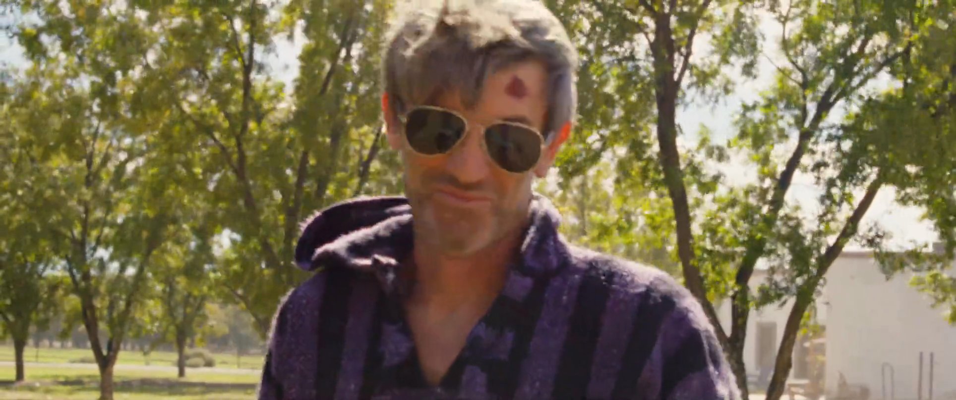 Two men engage in a tense, outdoor conversation amidst a sparse, arid landscape. One man, wearing a striped hoodie and sunglasses, gestures while speaking to his counterpart.