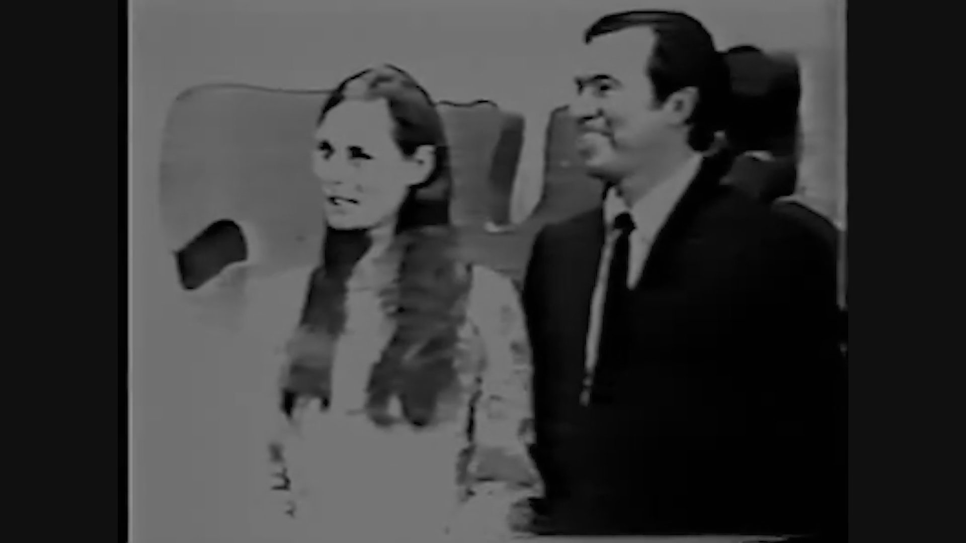 A woman participates in a televised game show conversation while a man provides commentary from off-camera. The black and white footage captures a candid exchange in a studio environment.