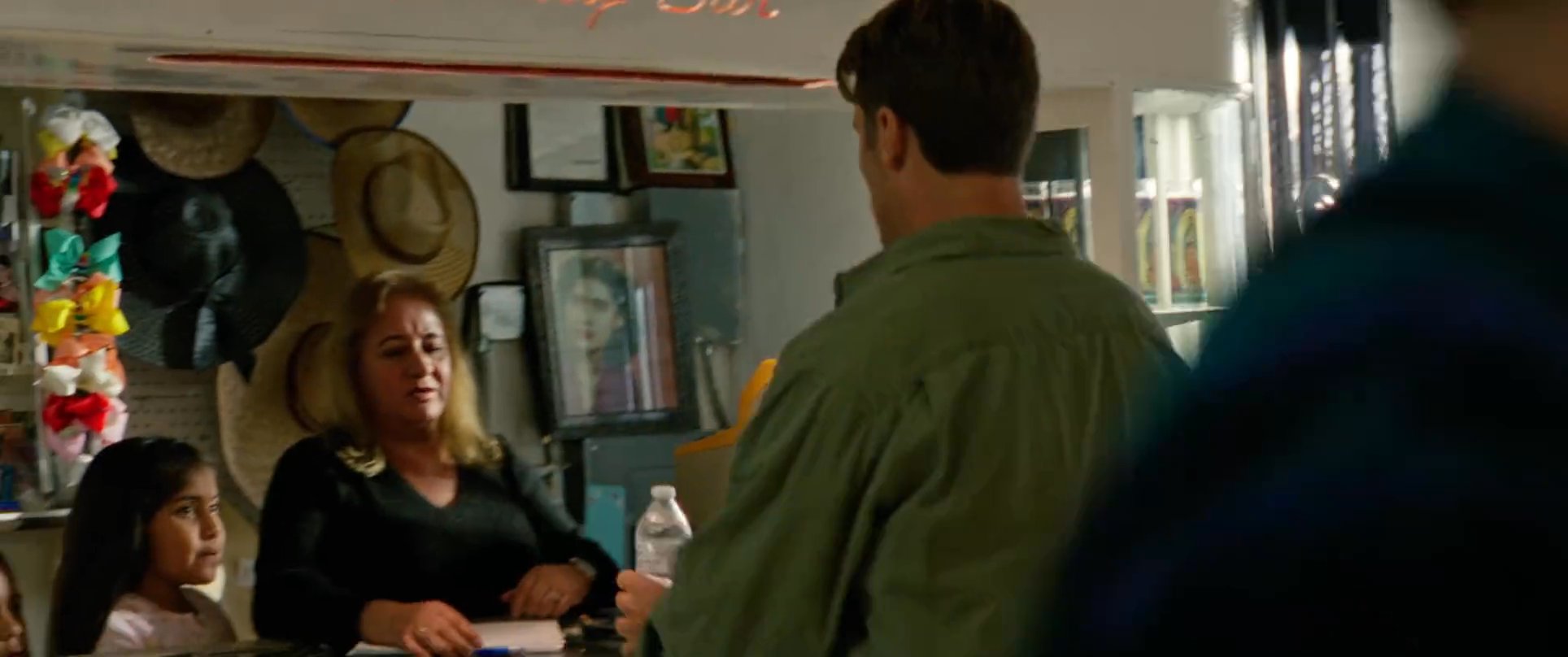 A man arrives by pickup truck at a small roadside store and completes a transaction at the counter with the clerk. The encounter takes place in a modest, quiet commercial space featuring a mix of retail goods.
