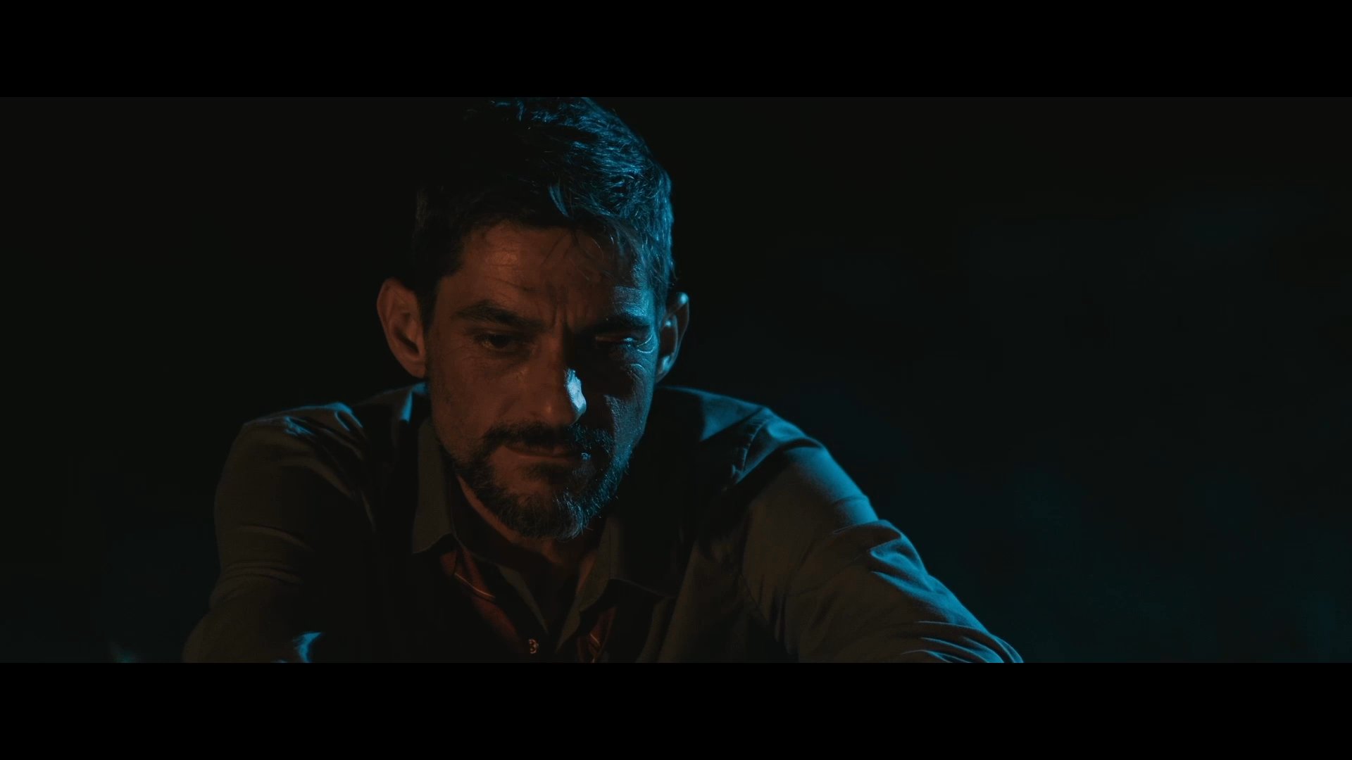 Two men engage in an intimate, low-light conversation where one recounts a personal failure regarding his son's school performance. The setting conveys a somber, reflective atmosphere as he shares his experience.