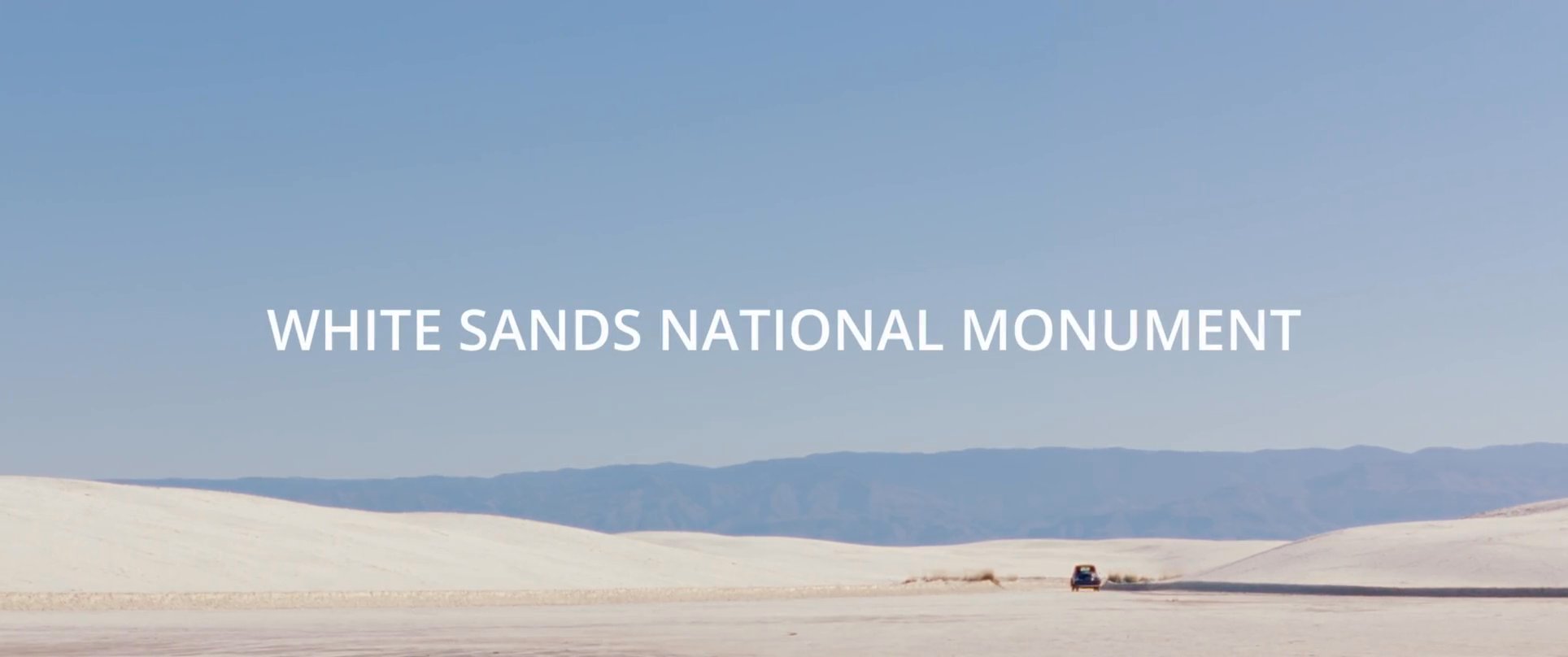 A vehicle drives across the expansive white gypsum dunes at White Sands National Monument under a clear, bright sky.