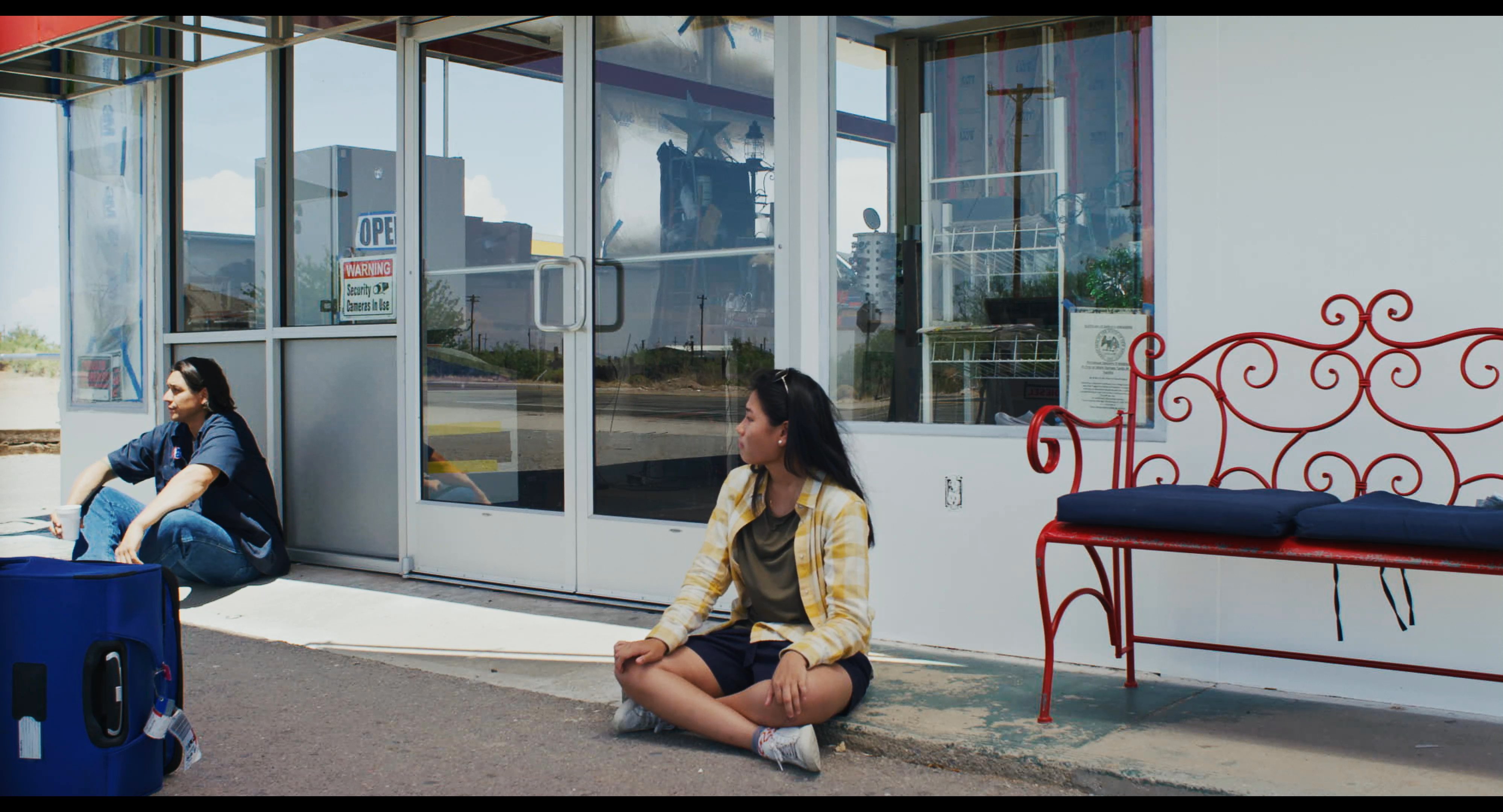 A woman sits in a meditative pose on a sidewalk outside a storefront while waiting with her luggage. She remains still and composed in a quiet outdoor setting.