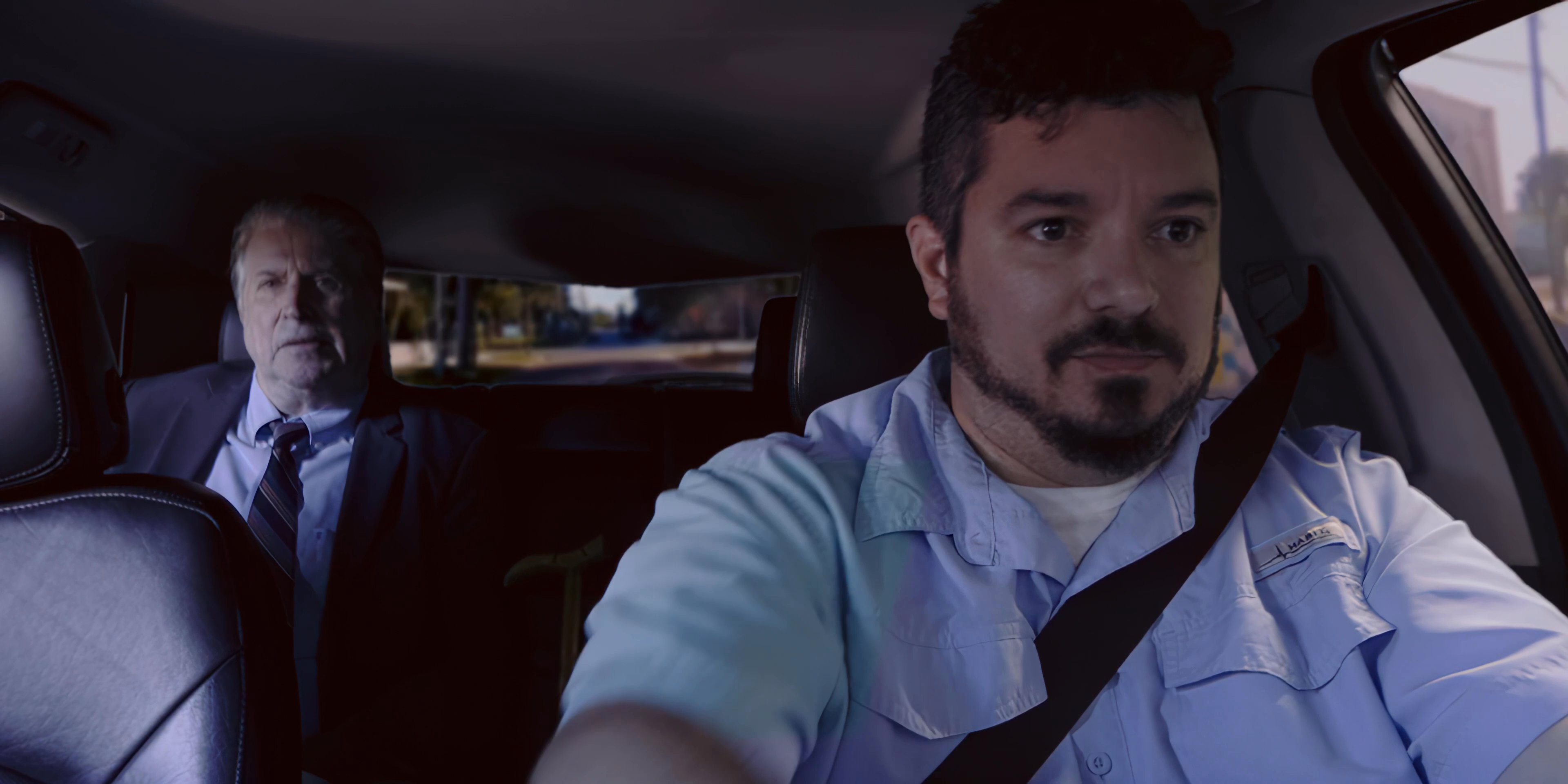 A driver and his passenger engage in a discussion while traveling through an urban environment. The two men maintain a serious demeanor during their commute.