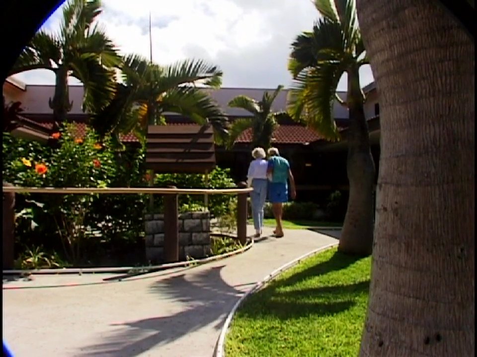 An elderly couple converses in a sunny, tropical outdoor setting before walking together along a garden path. The scene transitions to a surfer riding a wave in the ocean.