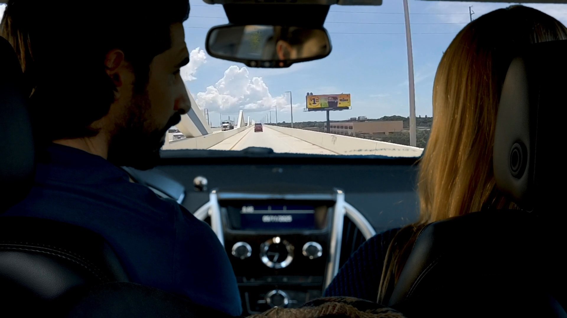 A man and woman share a conversation and hold hands while traveling together along an open highway in a vehicle.