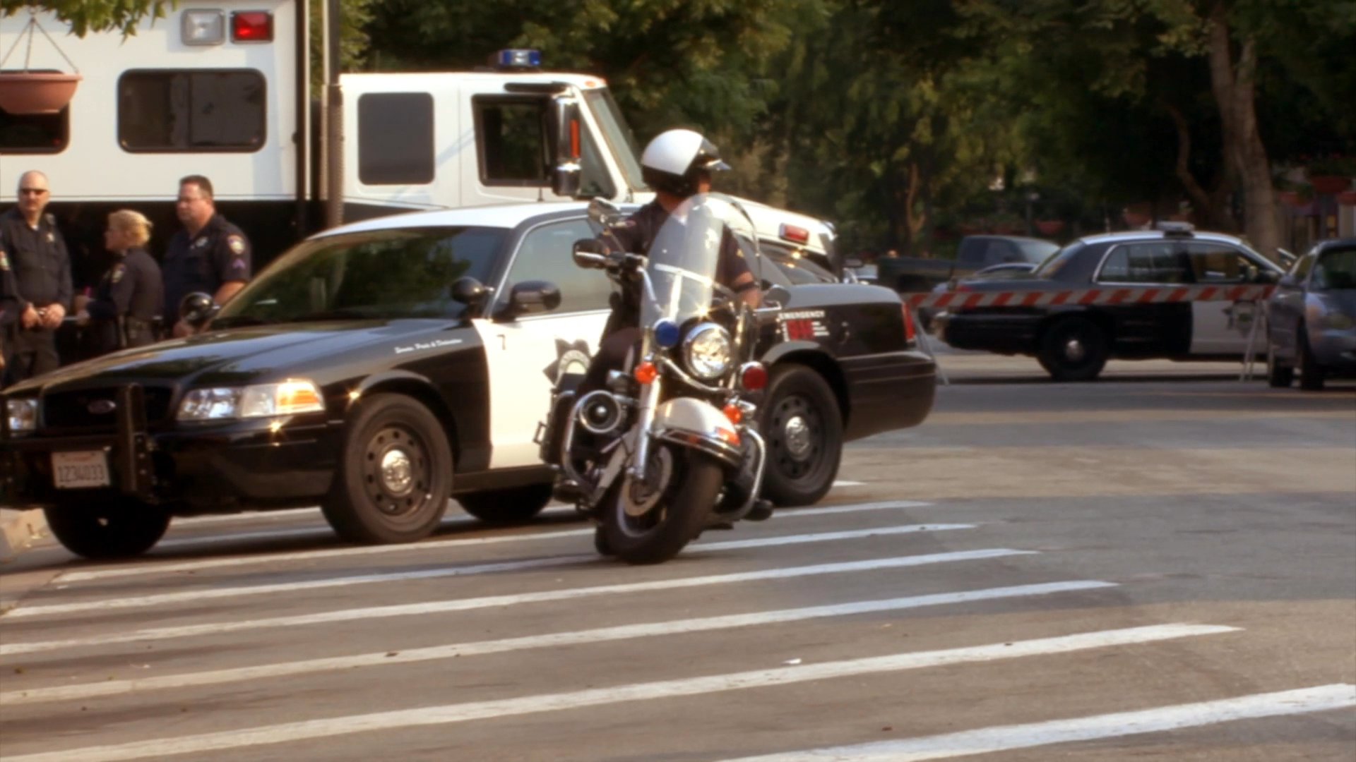 A man wearing a motorcycle club cap provides commentary while police officers manage traffic and monitor an intersection in a California town.