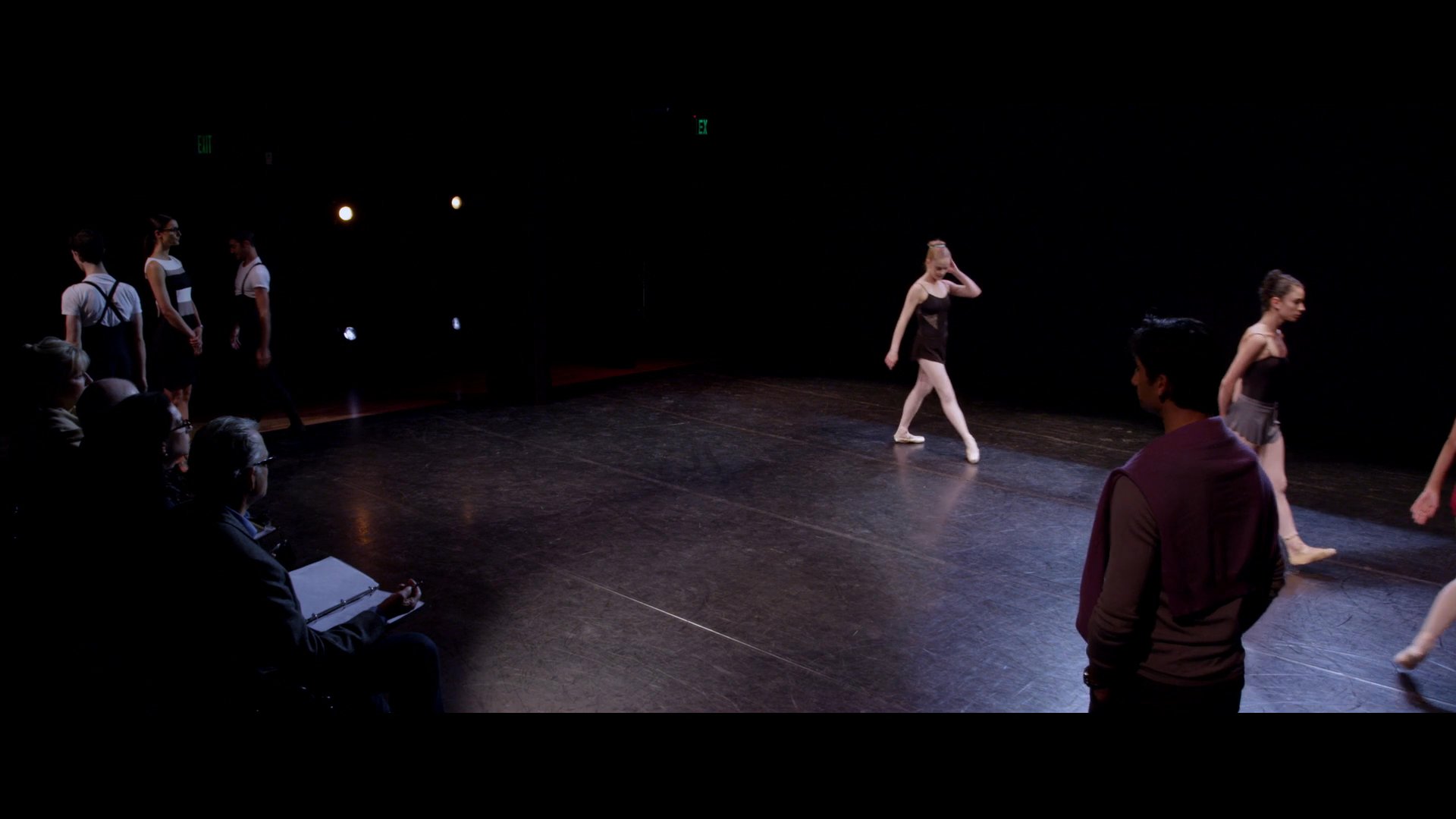 Dancers perform technical sequences on a stage during an audition while a panel observes. The focus shifts to a solo female dancer demonstrating her poise and classical technique.