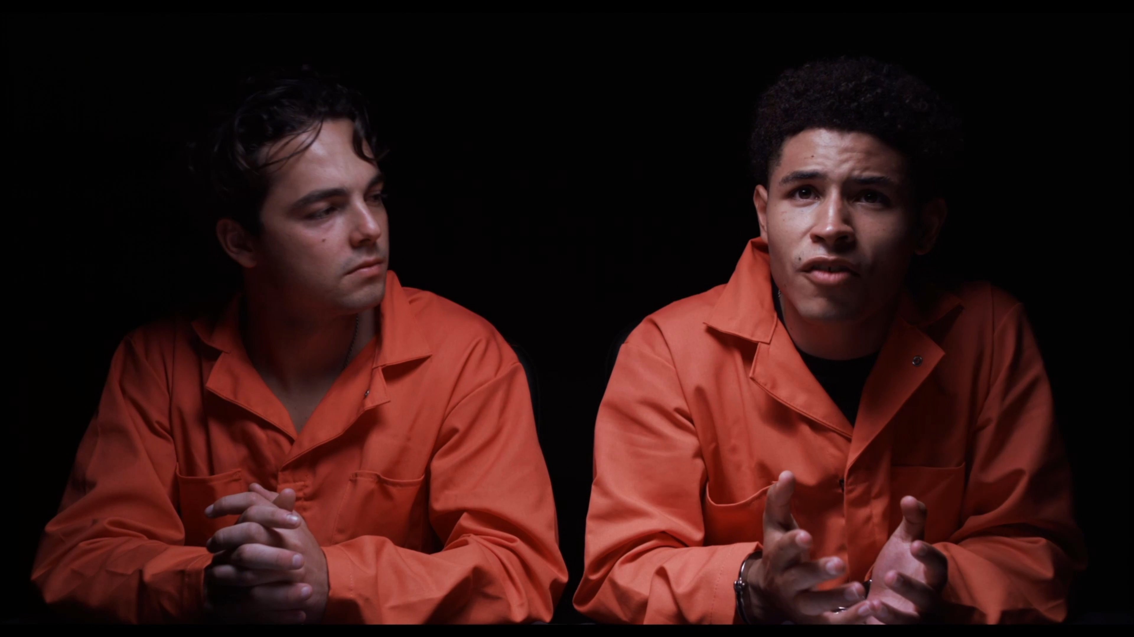 A federal agent in a DEA jacket conducts a formal interview while two individuals in prison jumpsuits listen and respond. The scene takes place against a dark background, focusing on the professional inquiry and the dialogue between the parties.