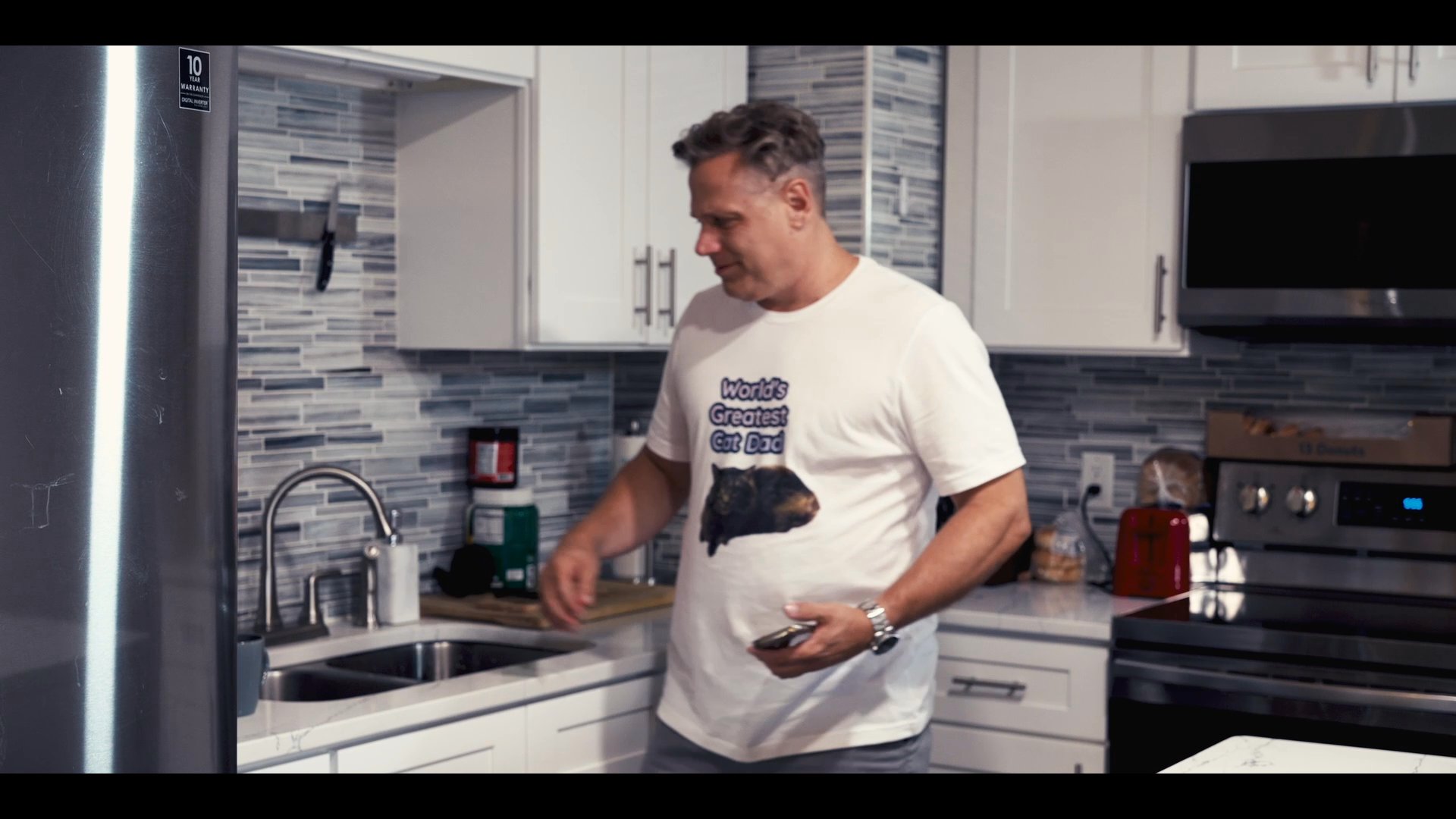 A man in a graphic t-shirt prepares coffee and checks his smartphone in a modern kitchen before the scene transitions to an aerial view of a suburban neighborhood.