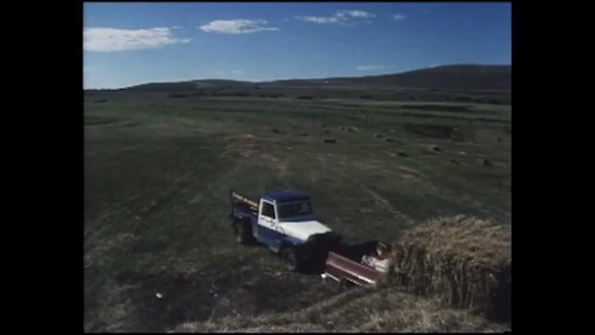 A farm worker in a cowboy hat walks among hay bales beside a pickup in a wide-open field, then speaks about the difficulty of finding people who can keep stacking hay load after load.