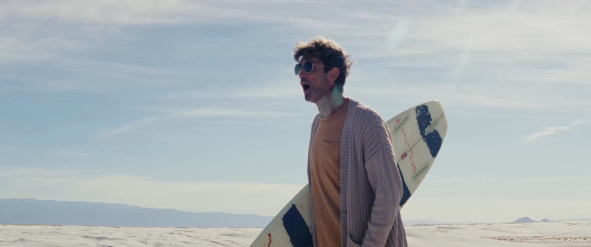 Two individuals navigate a vast, white sand dune while one person attempts a sandboarding run, resulting in a tumble. The scene captures the challenging terrain and leisure activity in a desert environment.