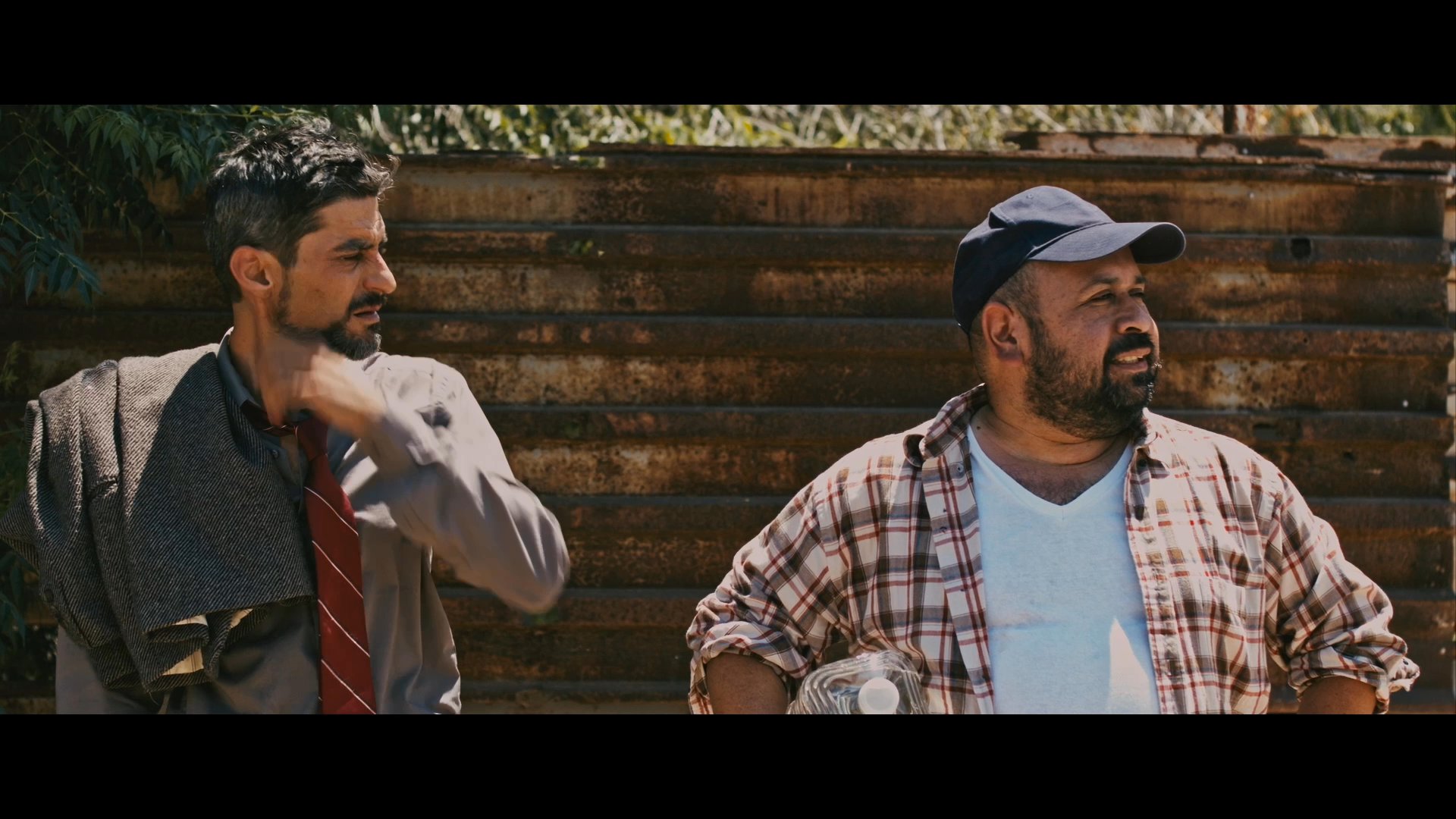 Two men have a tense conversation in a desert setting near a large border wall. The individual in the suit insists on departing today, while his companion expresses skepticism about the feasibility of the plan.