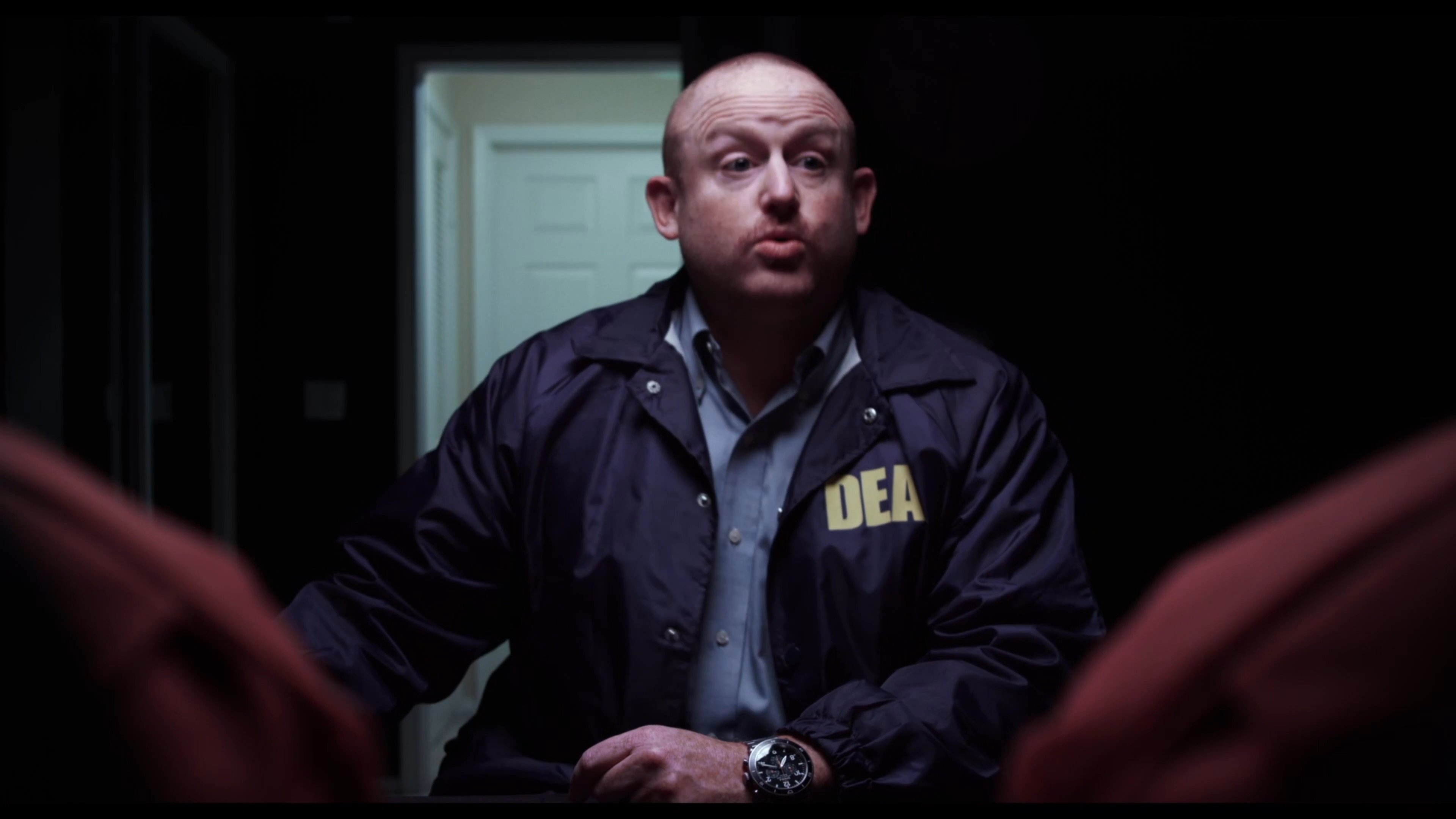A bald DEA agent in professional attire speaks while holding a striped coffee mug in a dimly lit, somber environment.