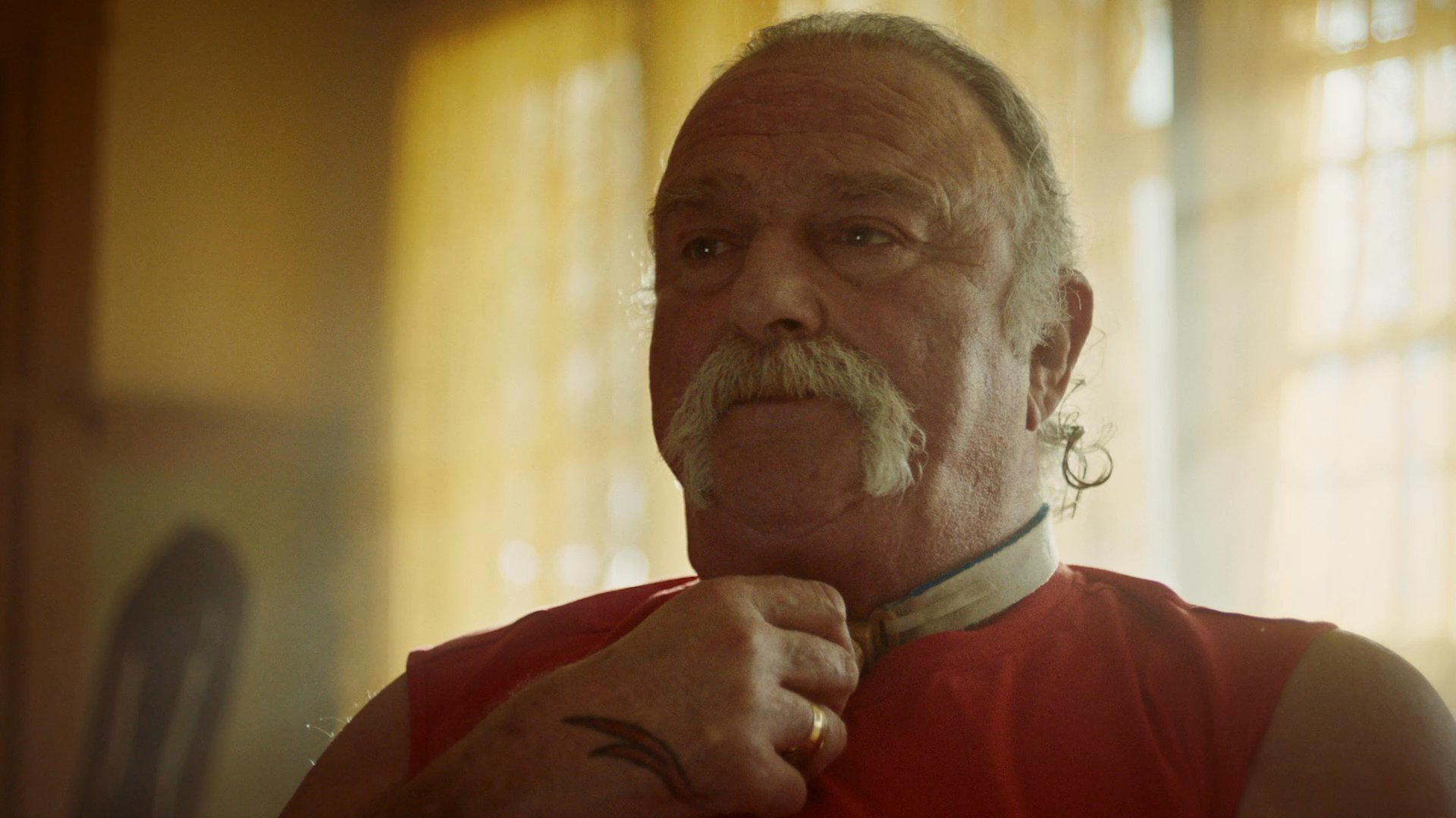 Two individuals sit on a couch in a sunlit living room while an older man with a mustache engages in a pensive discussion.