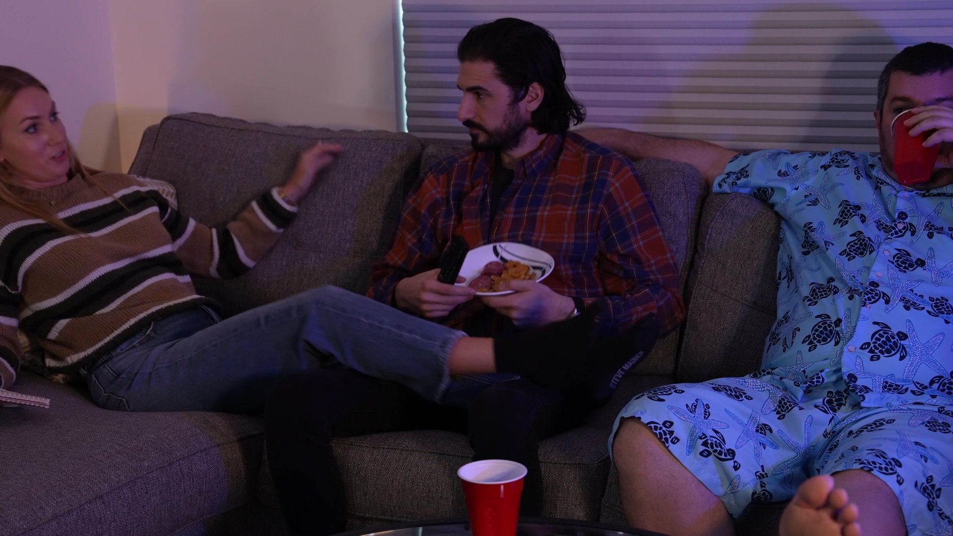 Three friends relax on a couch in a dimly lit living room, engaging in casual conversation and shared downtime.