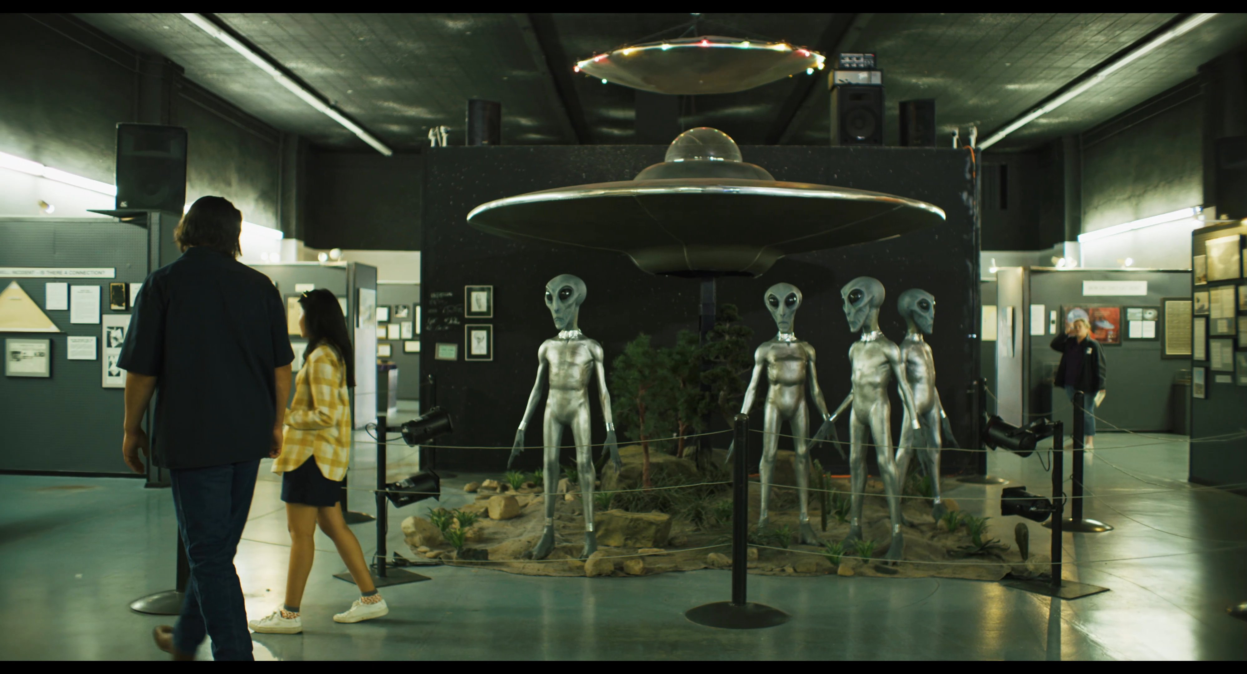 Two visitors walk through a museum gallery dedicated to extraterrestrial life, observing models of aliens and a ufo display.