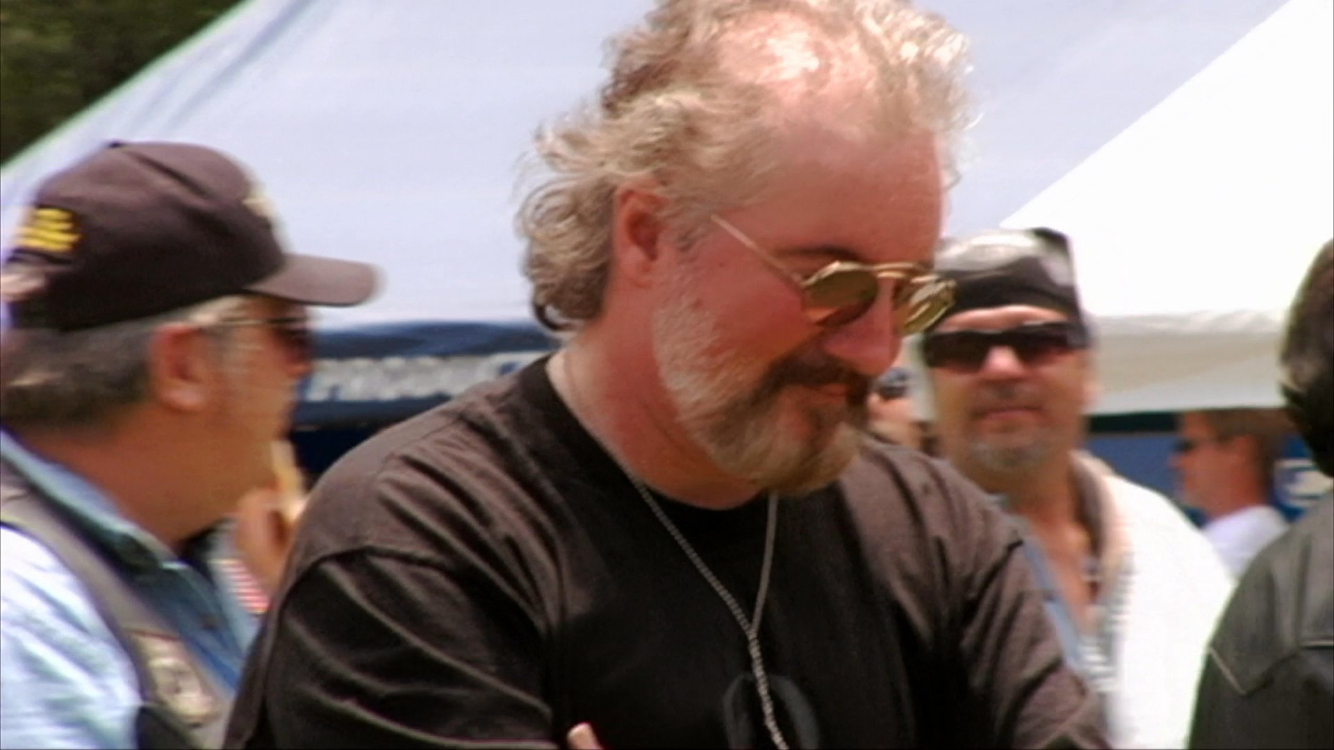 A rock band performs on an outdoor stage at a community event, featuring a bearded lead singer and musical accompaniment. The scene transitions to shots of the engaged audience members watching the live show.