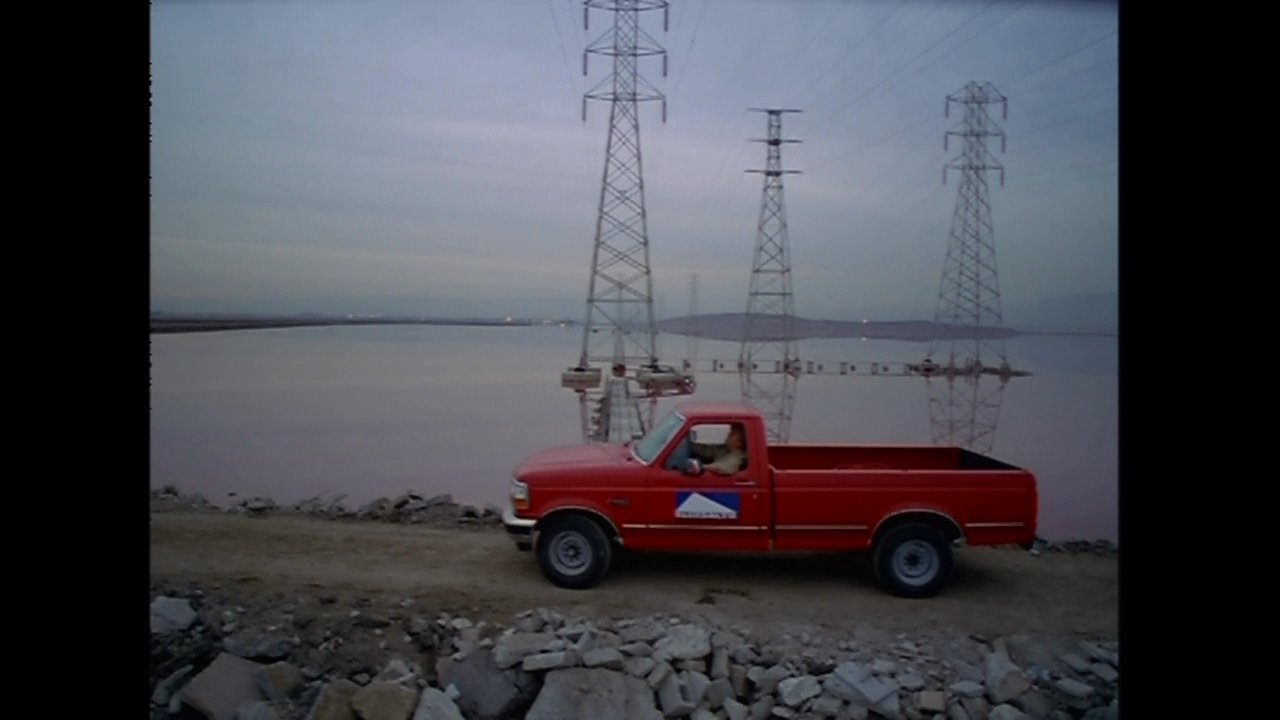 Two individuals engage in a conversation inside a brightly lit eatery before transitioning to a scenic outdoor landscape featuring a red pickup truck driving along a shoreline at dusk.