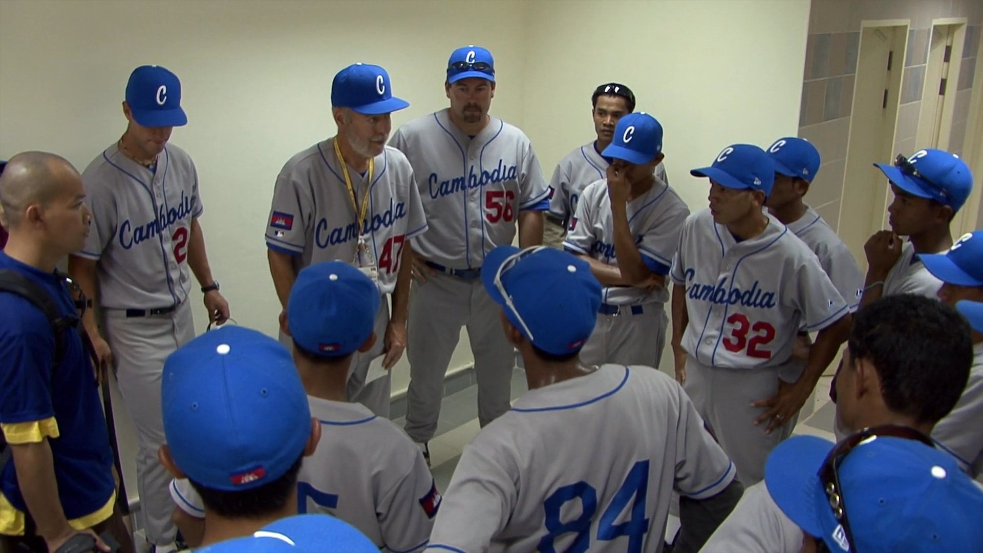 A baseball team gathers in a locker room for a focused pregame huddle before taking the field for a match against Thailand. The players and coaches exchange final instructions and rally together to build team morale.