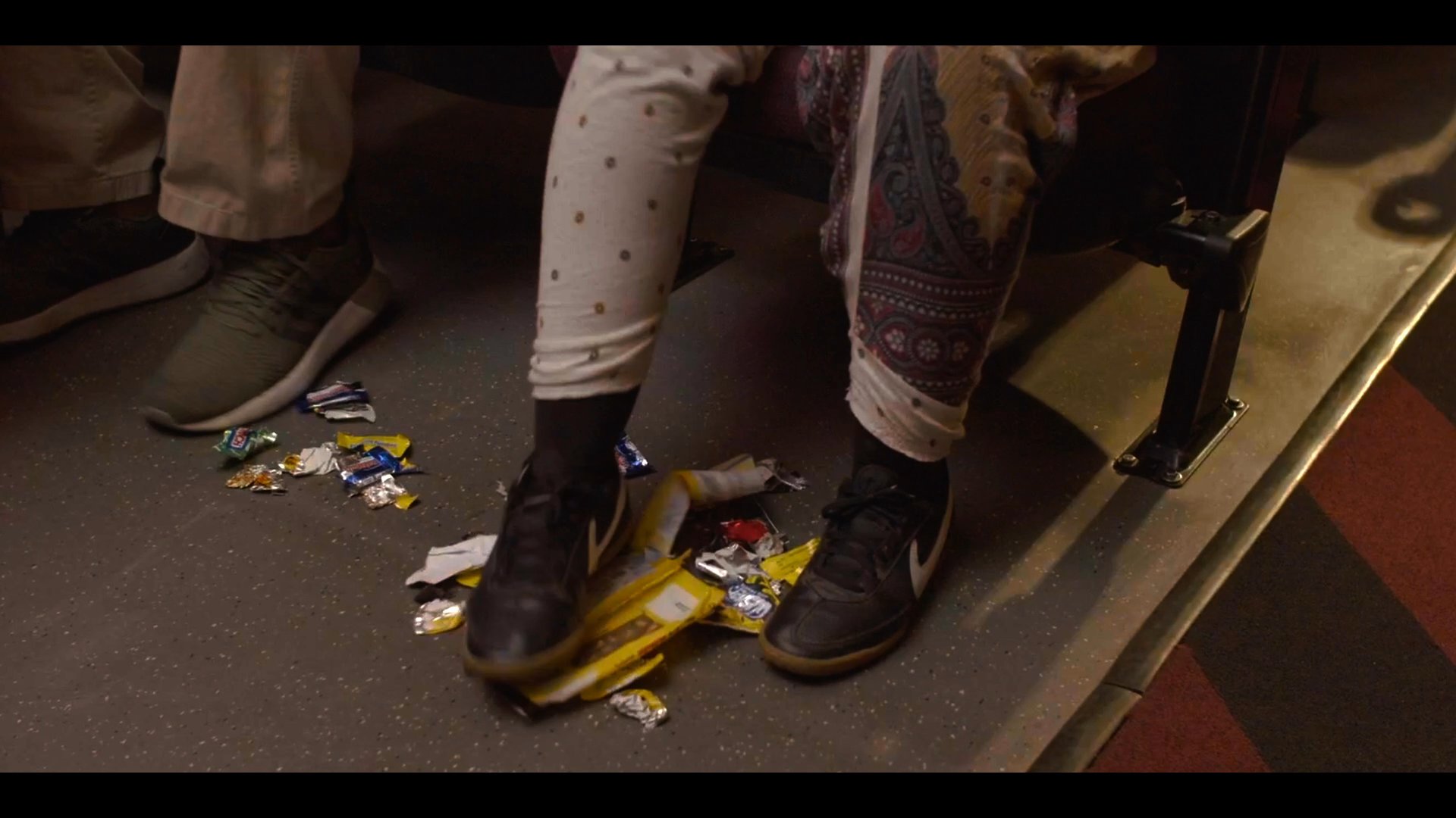 Two individuals consume snacks inside a movie theater, leaving significant amounts of trash on the floor beneath their seats before departing. The sequence captures their casual disregard for the space as they exit the auditorium.
