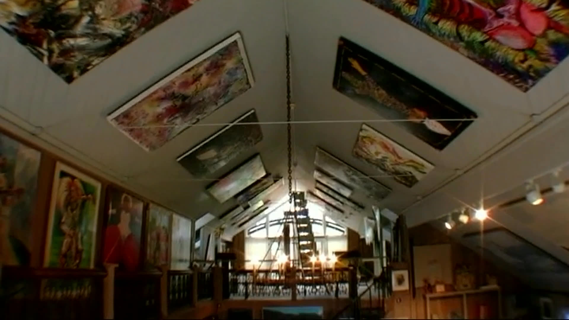 A crowded, multi-level home is filled with paintings, sculptures, and collected objects, with a dining table at its center. The speaker recalls years of family dinners there and notes that the room once held fewer belongings.
