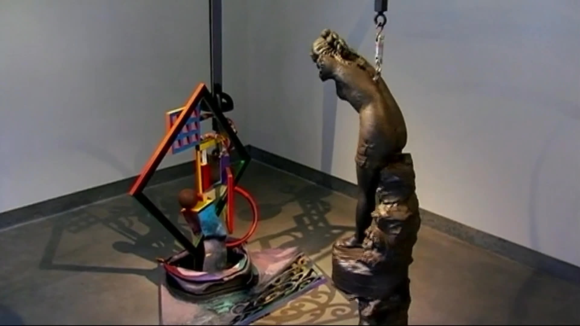 A woman speaks from a book-lined room while views of a rural landscape and unusual gallery artworks appear, including a colorful assemblage and dark sculptural forms. She recalls a piece she calls the “battle house” and says Renee’s genuine enthusiasm for what he buys makes her happy.