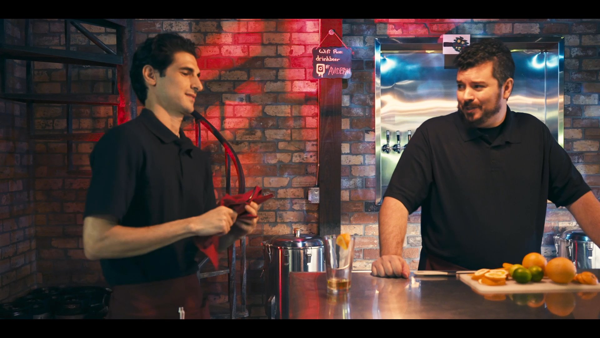 A bartender practices service techniques with a colleague at a brewery bar while customers relax in the background. The staff focus on professional presentation and engagement within the industrial-style establishment.