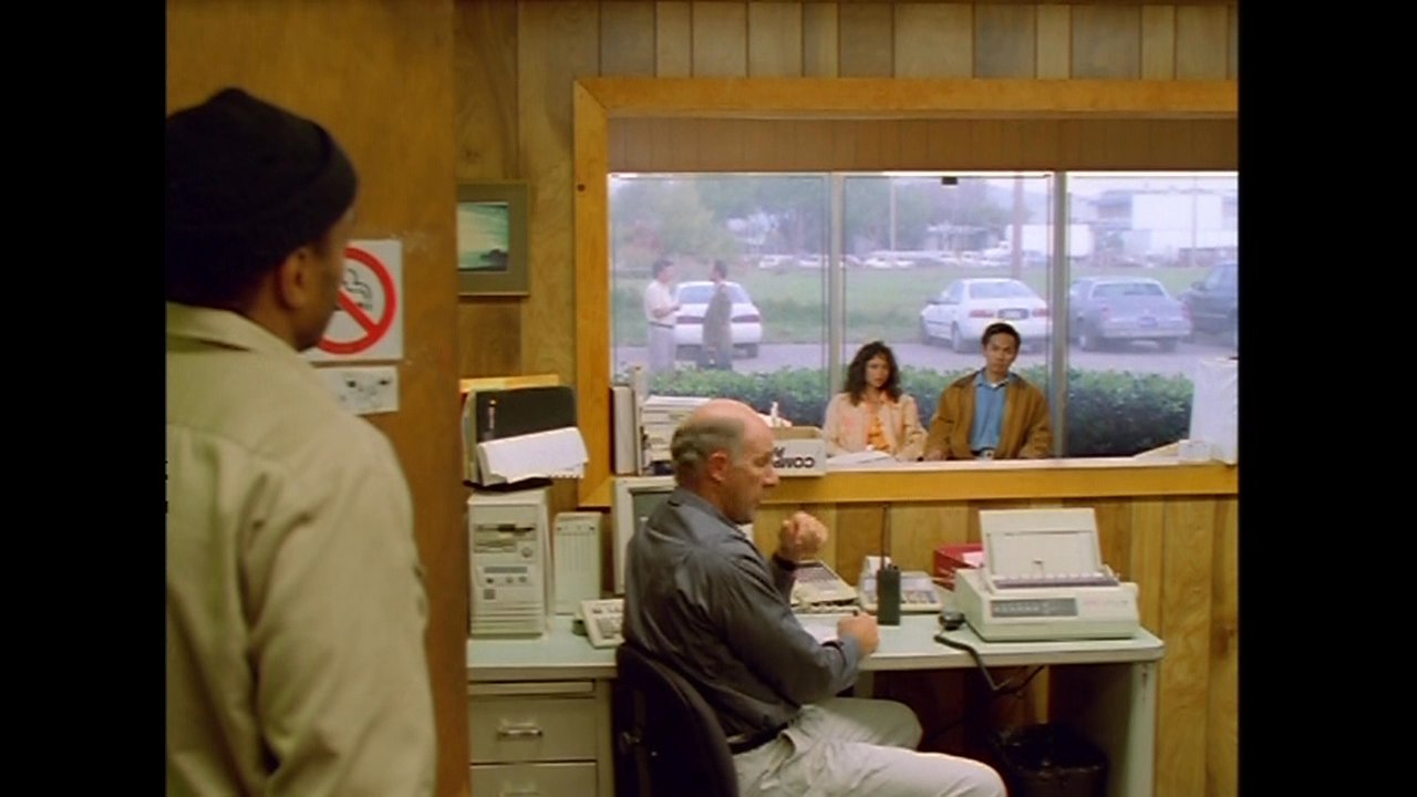 A man works in a wooden-paneled office overlooking a large salt processing site, while visitors wait in an adjacent lobby area.