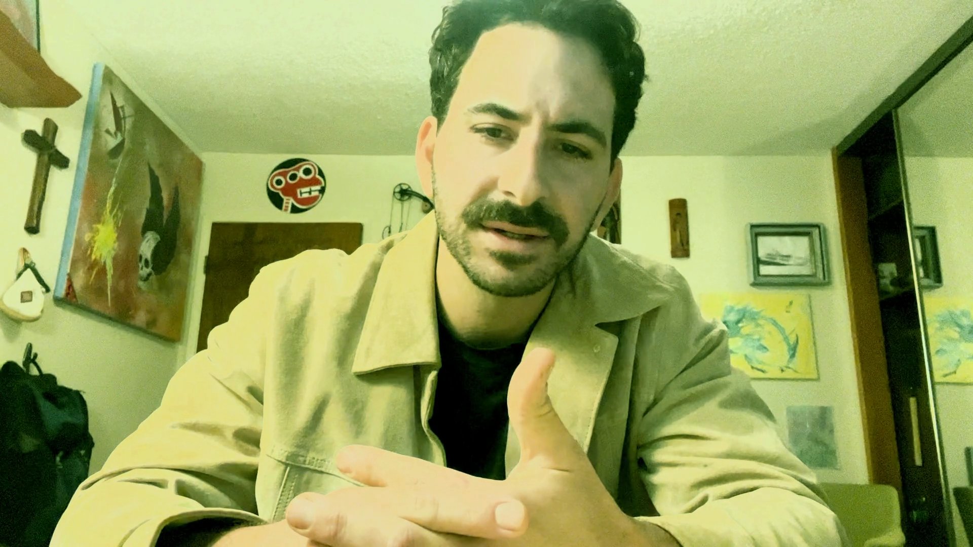 A man sits at a table and speaks directly to the camera, engaging in a focused and expressive monologue within an indoor setting.