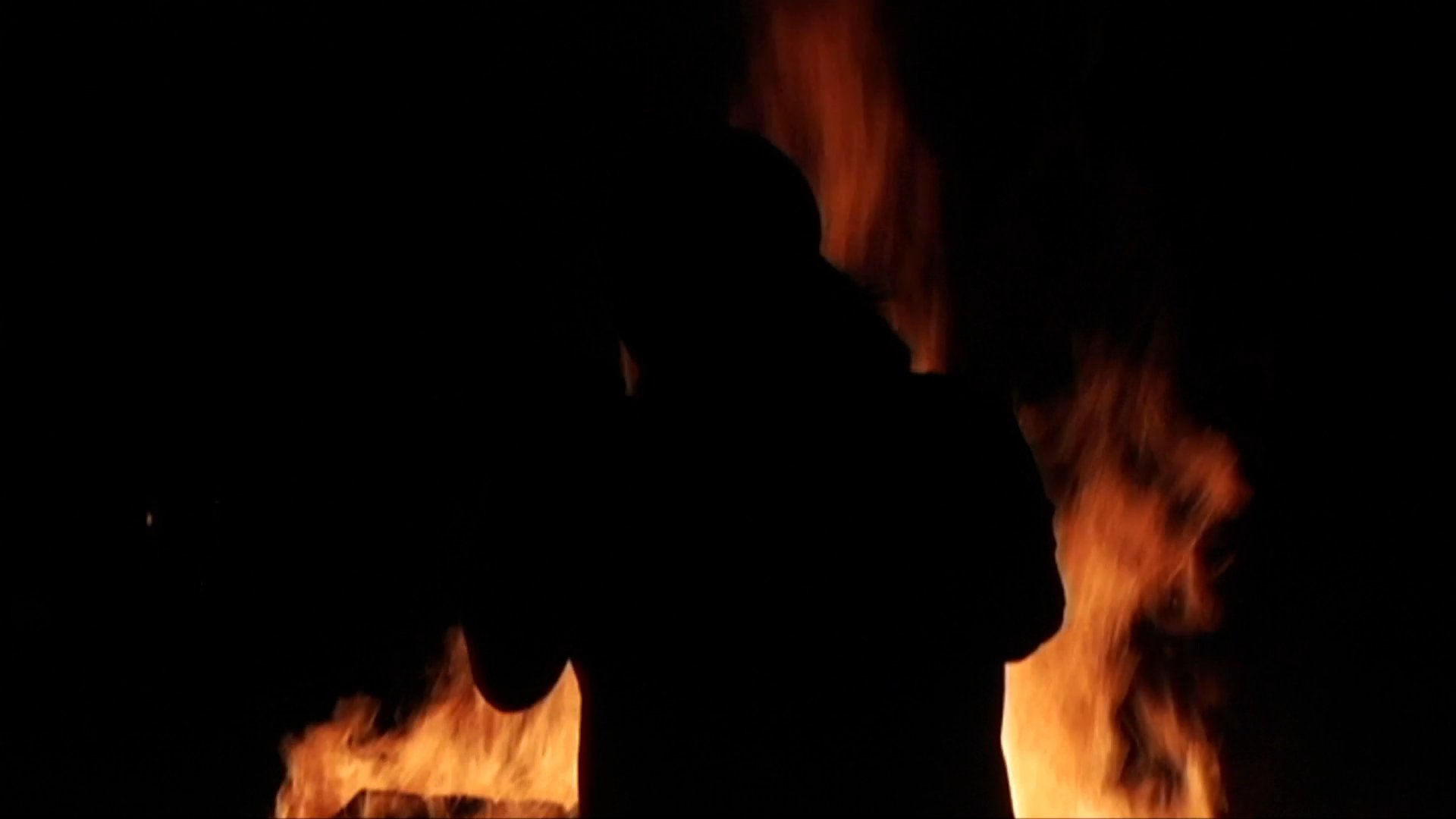 An artist performs in silhouette against an intense, blazing fire backdrop during a nighttime event. Dramatic lighting transitions from high-intensity lens flares to a dark, contrast-heavy atmosphere.