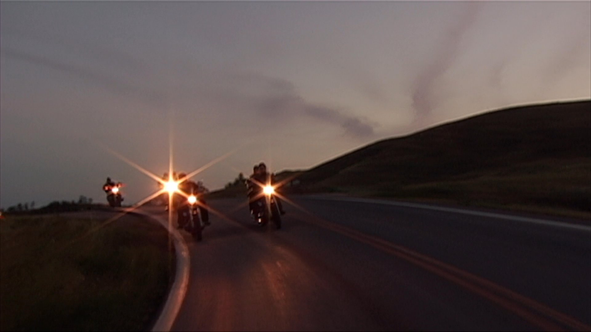 A group of motorcyclists cruises down a scenic highway during sunset, transitioning to a live indoor concert performance featuring a bearded vocalist and guitarist.