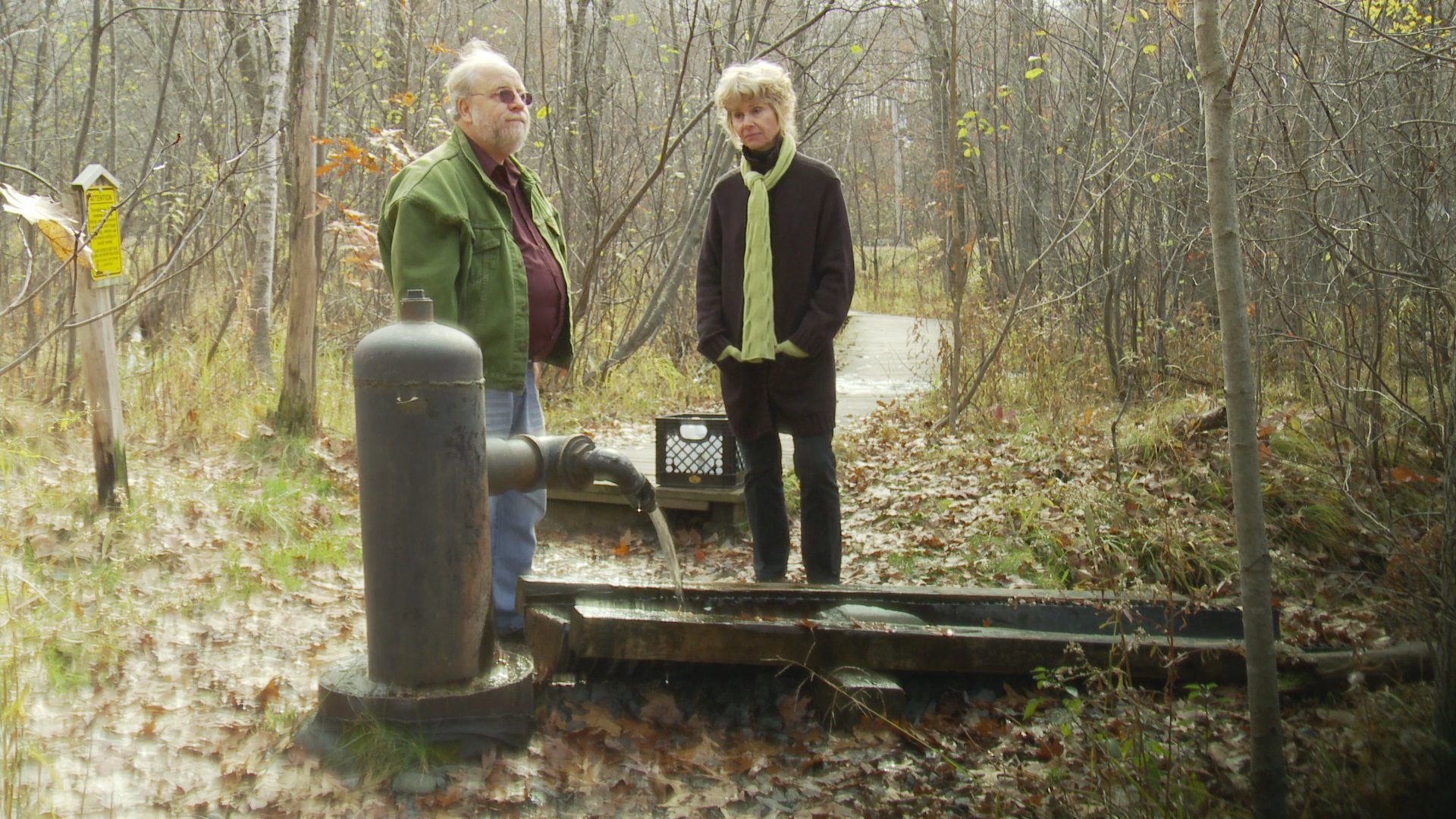 Two researchers conduct water quality sampling at a natural water source in a wooded area. The individuals collect a sample from an outdoor tap into a container as part of their monitoring process.