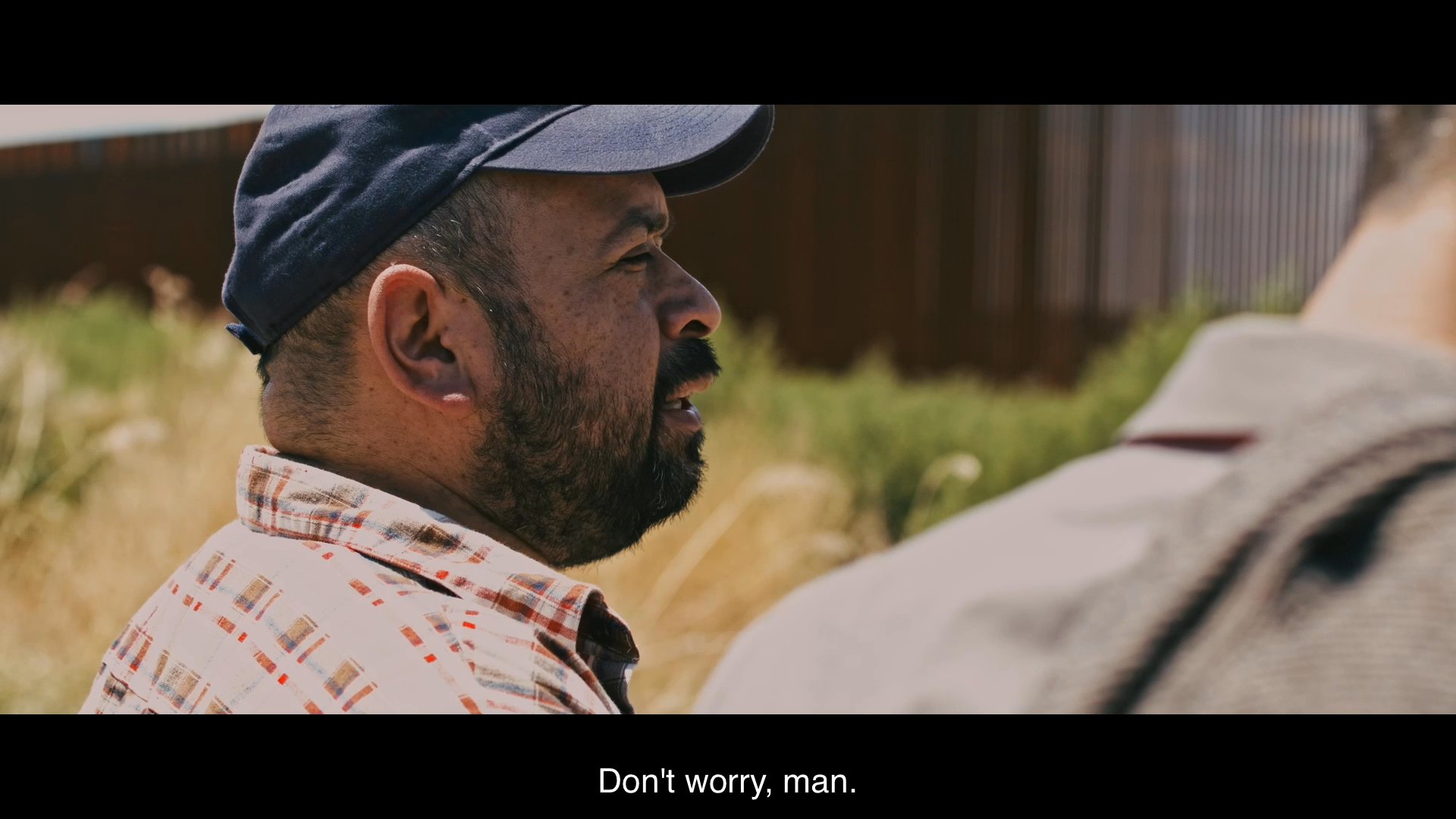 Two men stand near a border fence while discussing the duration and logistics of their journey. They share past experiences and plan an alternative route to continue their crossing.