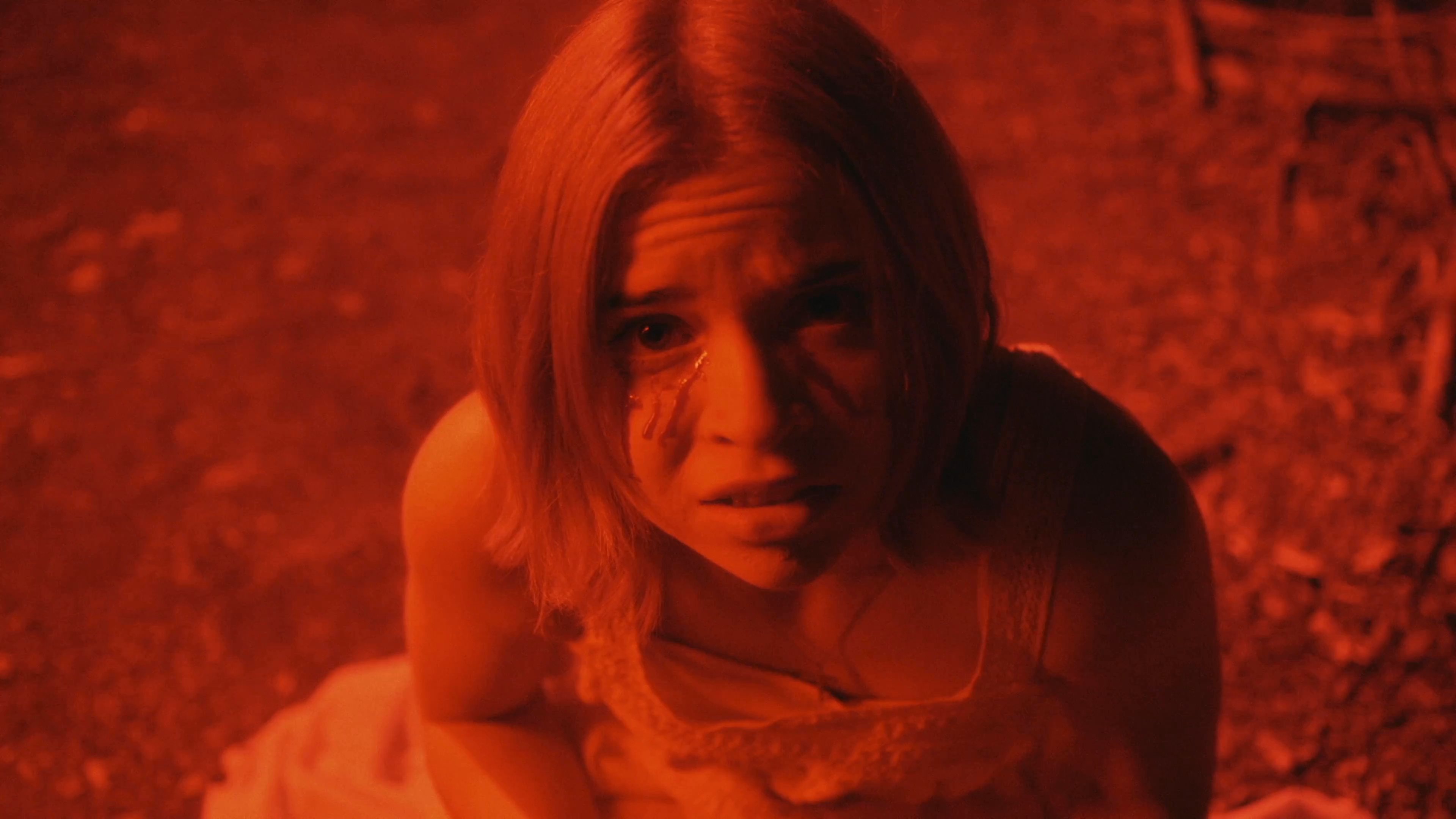 A woman in distress, marked by dark streaks on her face, kneels in a dimly lit, red-hued environment while confronting a mysterious figure wearing a floral crown. The atmosphere suggests a high-stakes, suspenseful encounter.