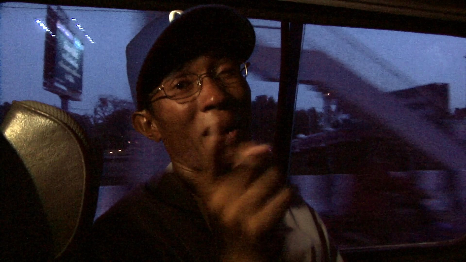 A passenger wearing a blue baseball cap sits in the back of a moving vehicle during twilight, looking out the window while subtly gesturing with his hands.