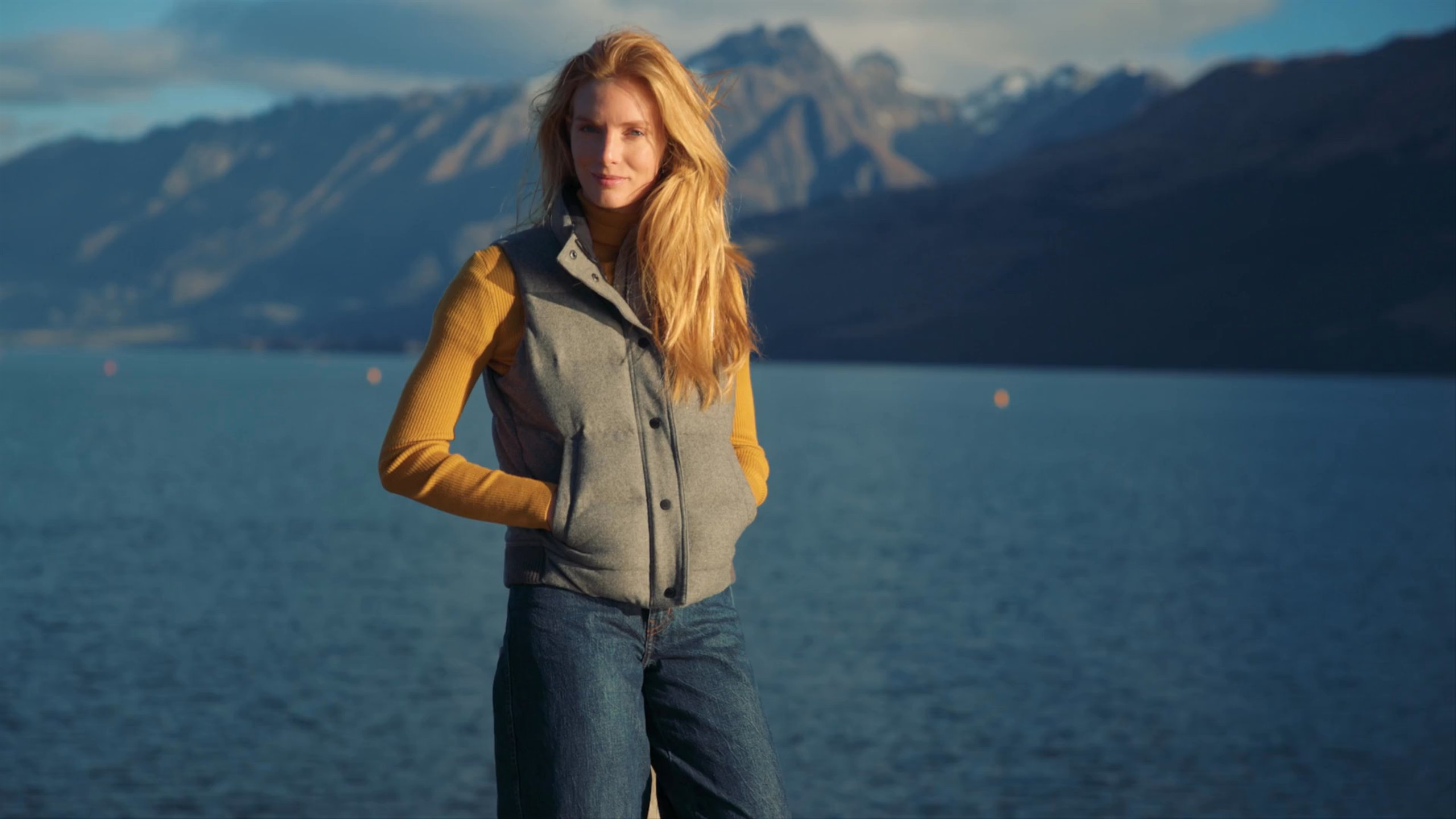 A woman stays and relaxes next to a lake in New Zealand and smiles down, with snow mountains in the background.