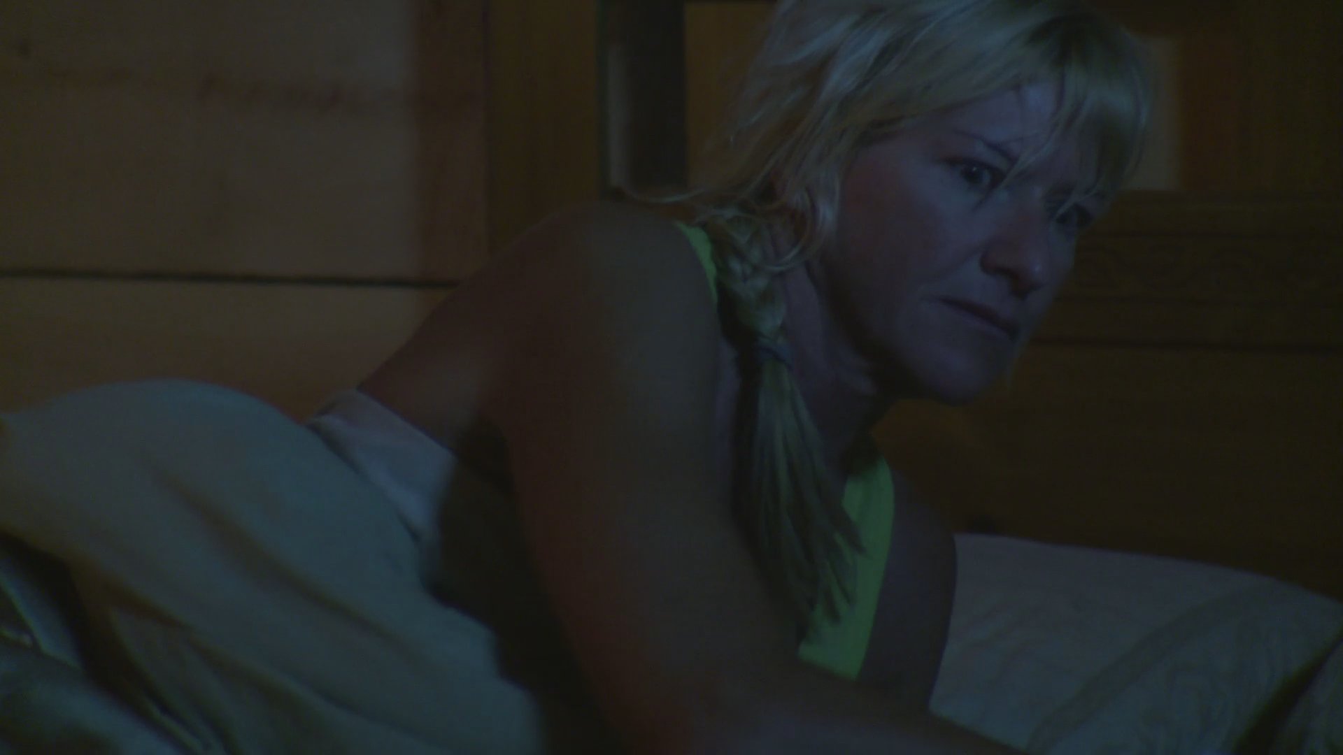 A woman wakes up suddenly during the night in a wooden bedroom cabin, appearing disoriented and distressed as she sits up in bed.