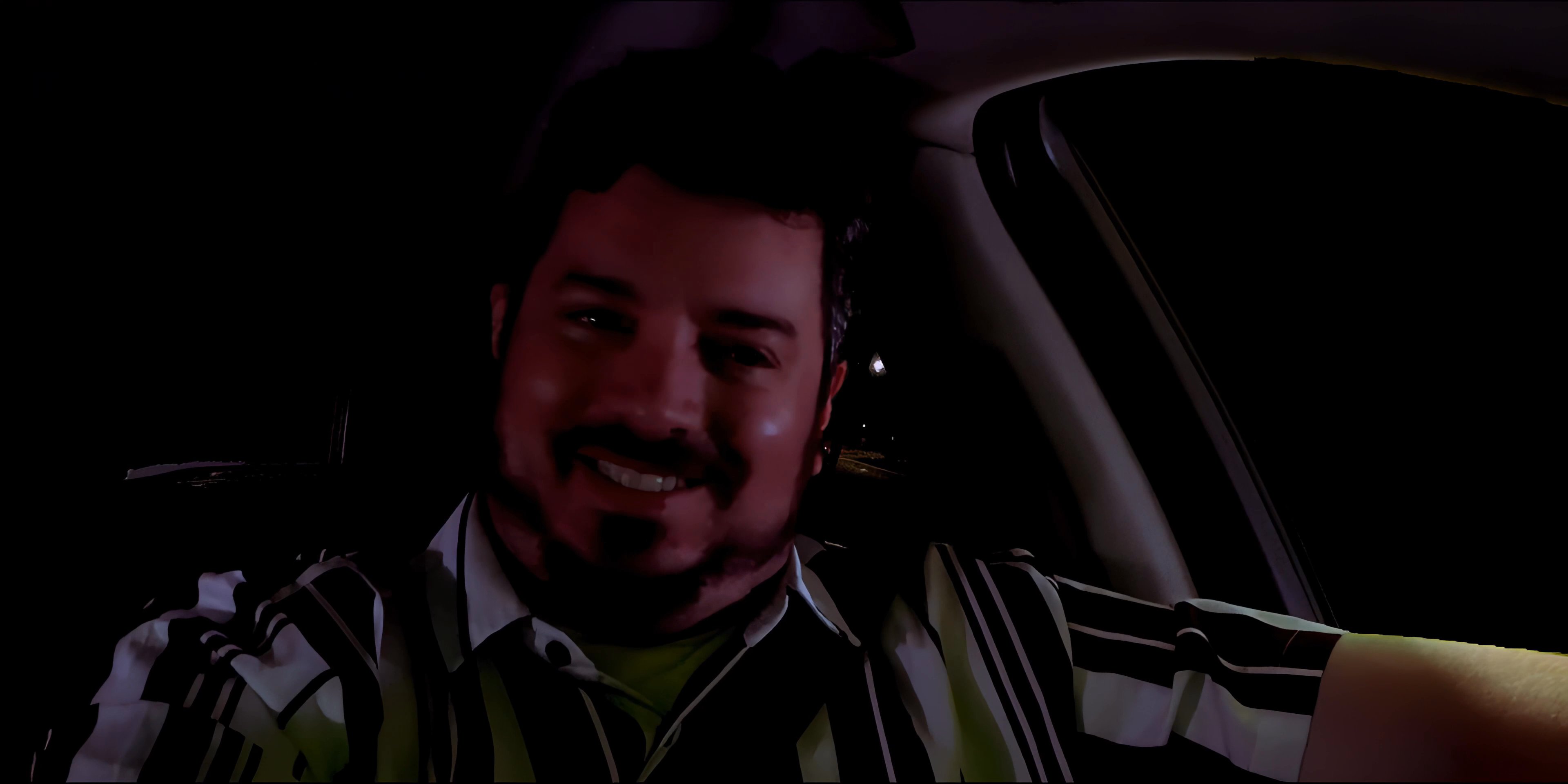 A man speaks to an unseen passenger while operating a vehicle on a dark road at night.