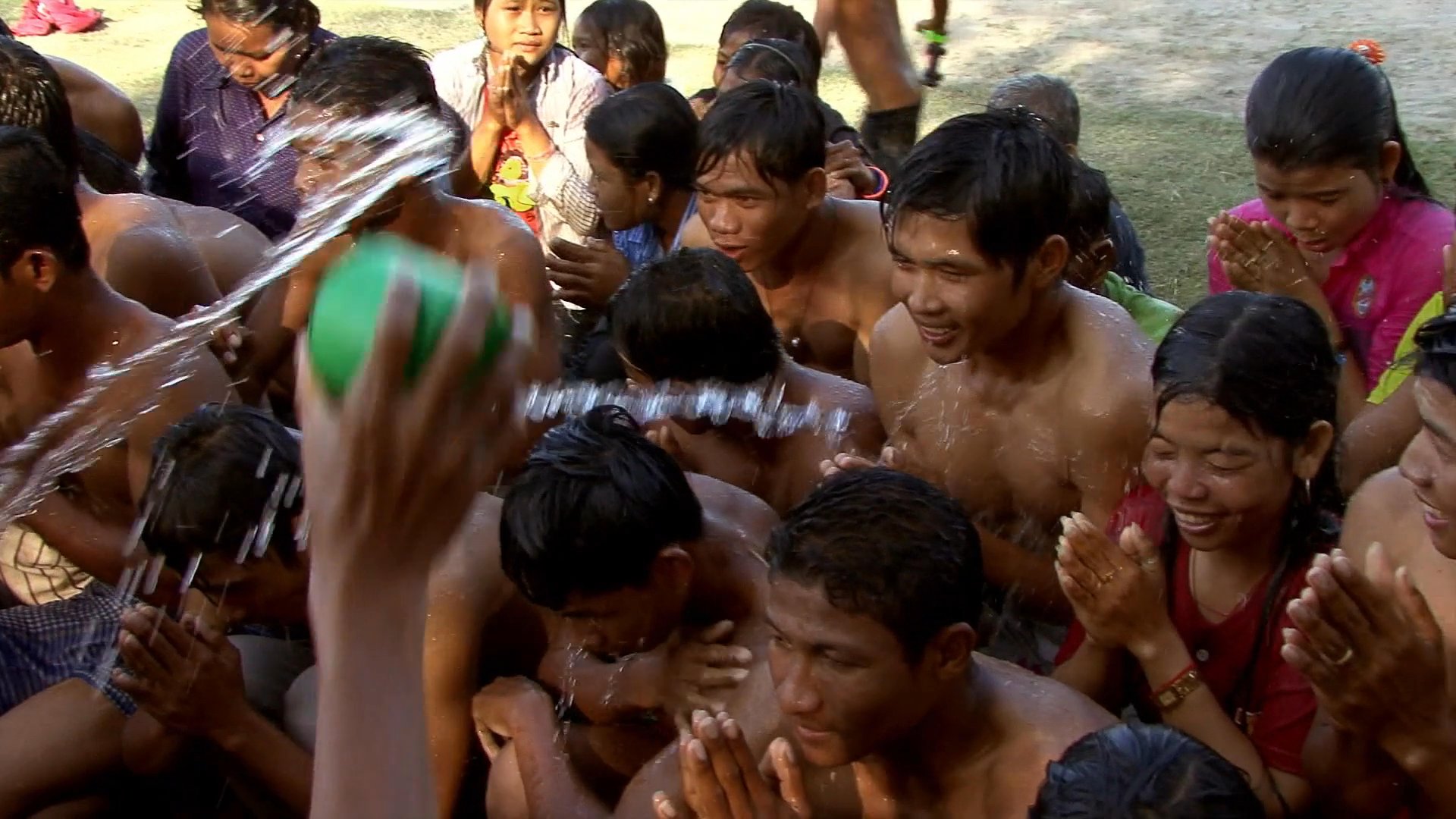 A Buddhist monk performs a blessing ritual by sprinkling water over a gathered congregation. Participants show reverence through prayer gestures as they receive the ritual water.