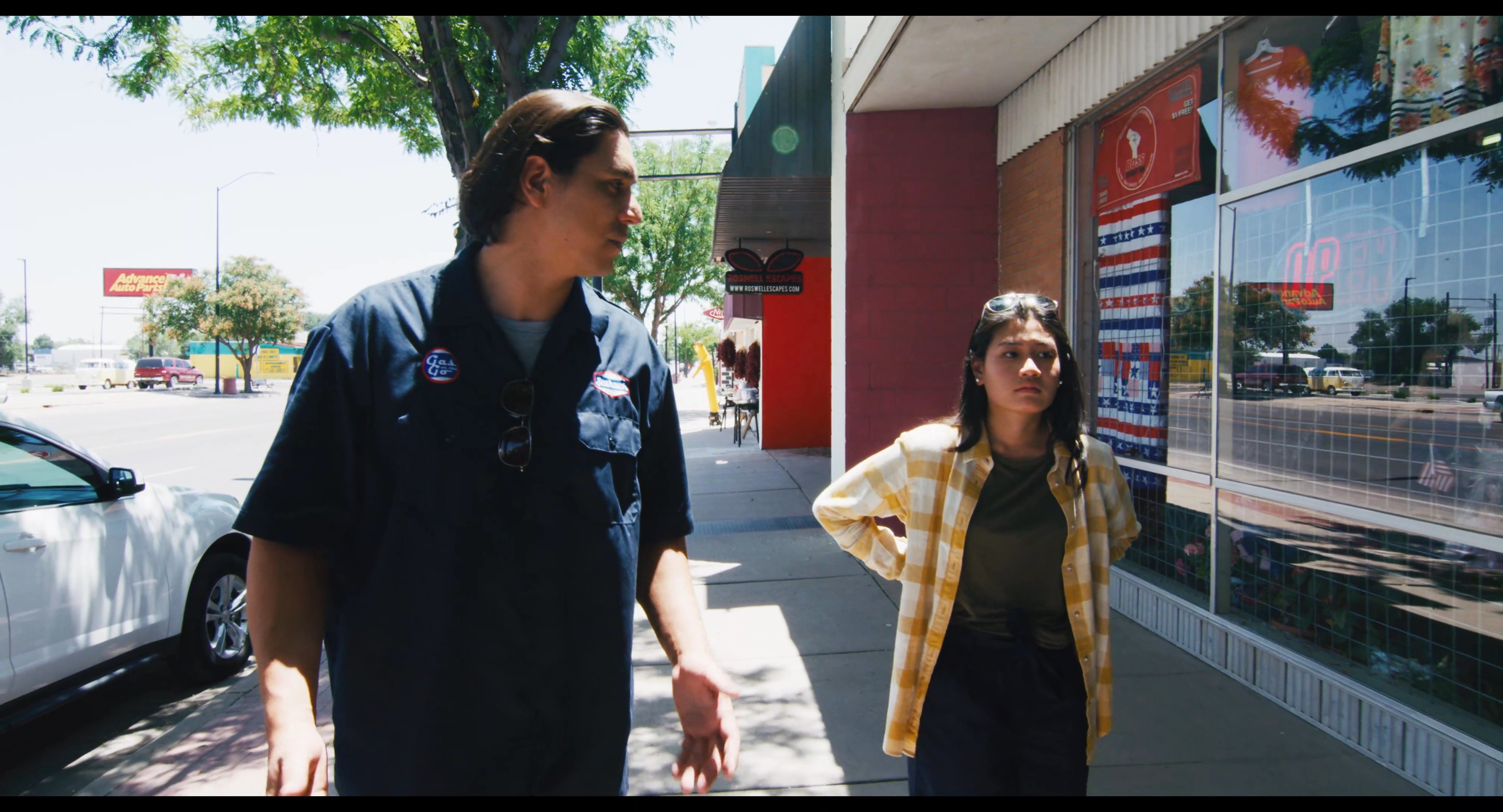 Two individuals engage in a discussion while walking along the exterior sidewalk of a commercial retail center. They maintain eye contact and gesture as they traverse the storefronts.