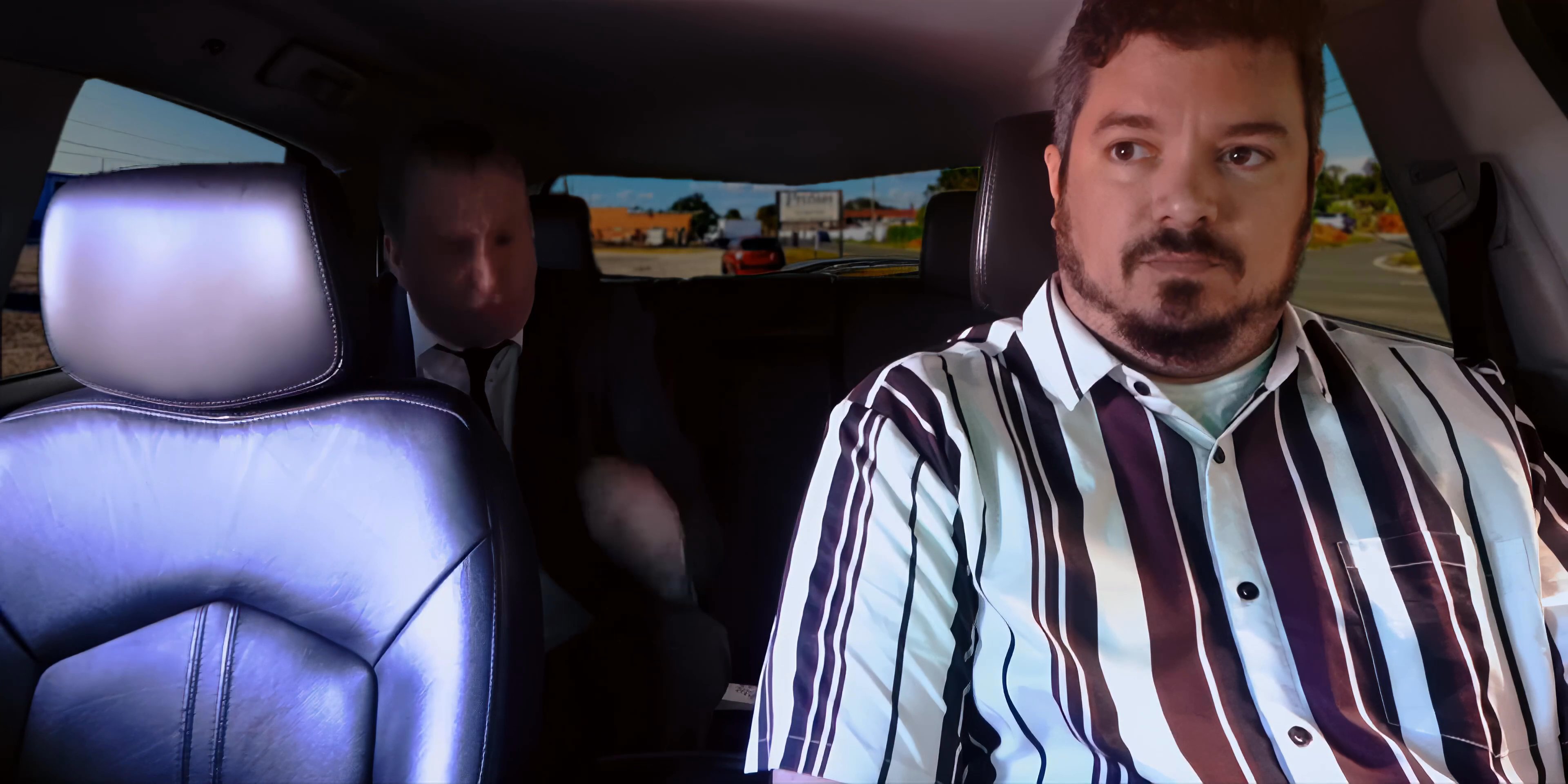 Two men engage in a serious conversation while traveling in the back of a vehicle, with one passenger visibly animated during the discussion.