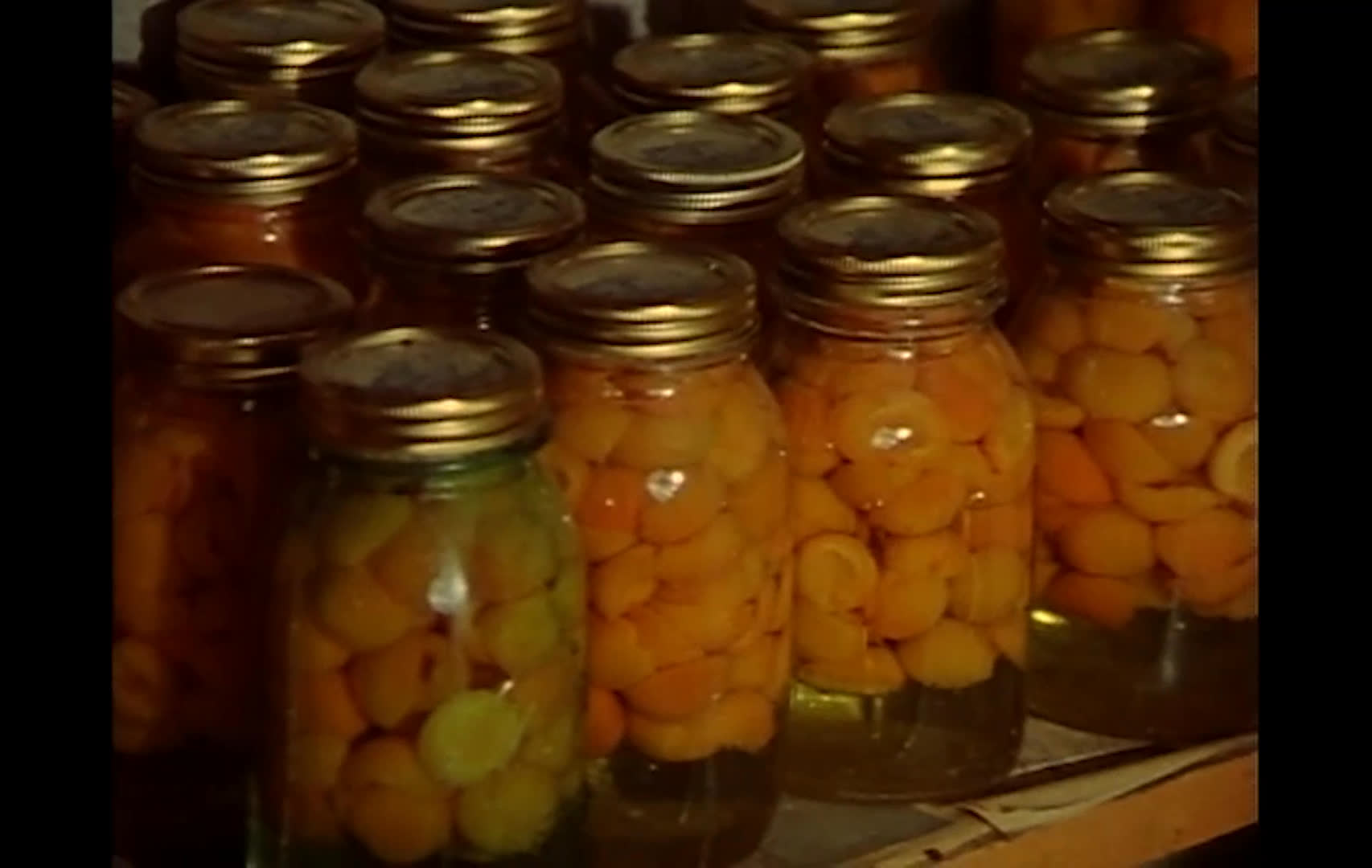 A man discusses a self-reliant household while standing beside shelves packed with jars of home-preserved vegetables and fruit. He describes canning the garden harvest, freezing peas, raising pigs, keeping eggs, and making butter to keep store costs low.
