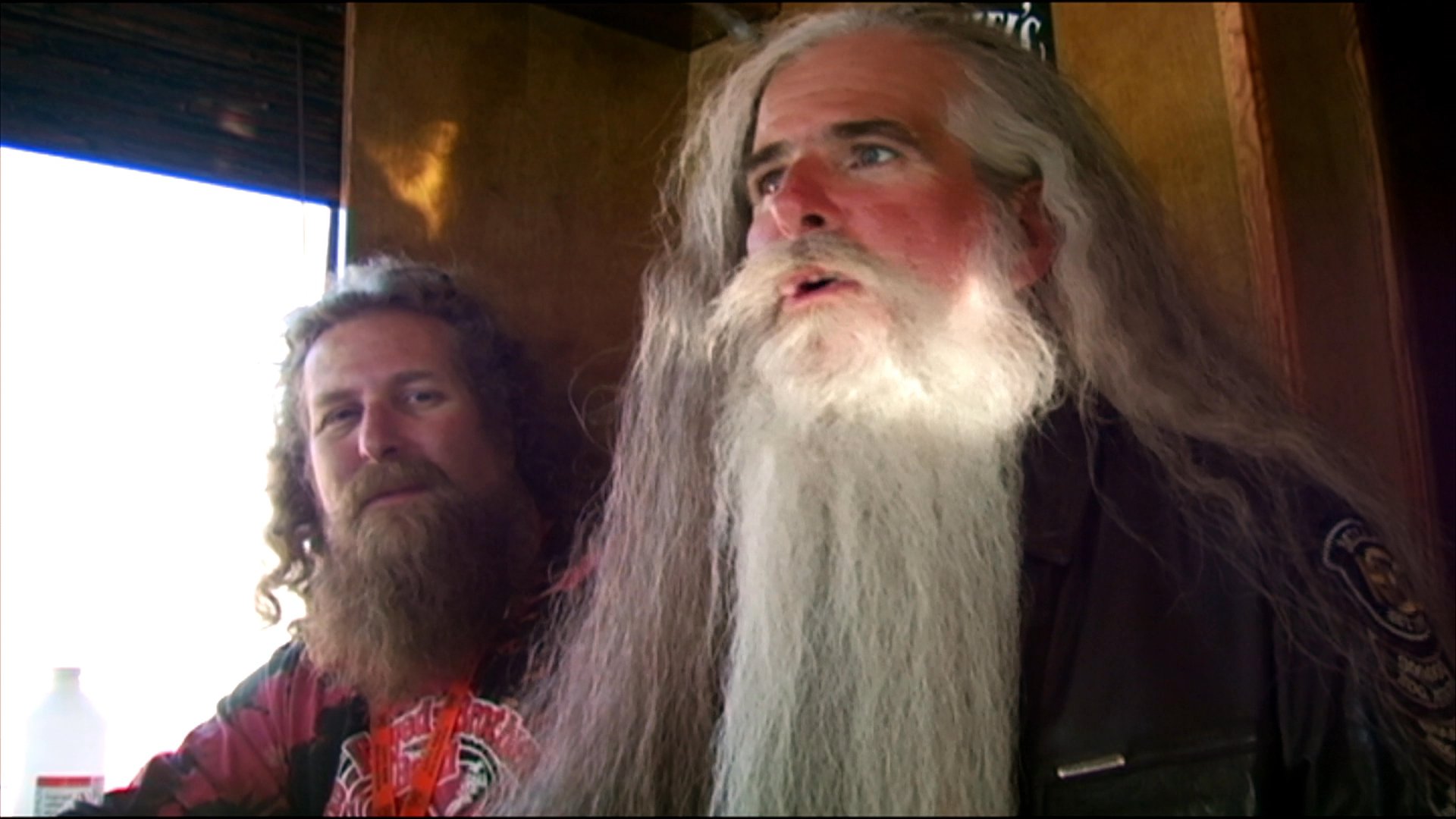 Two individuals with prominent facial hair discuss their experiences at a motorcycle rally while situated in a parking area filled with numerous motorcycles.