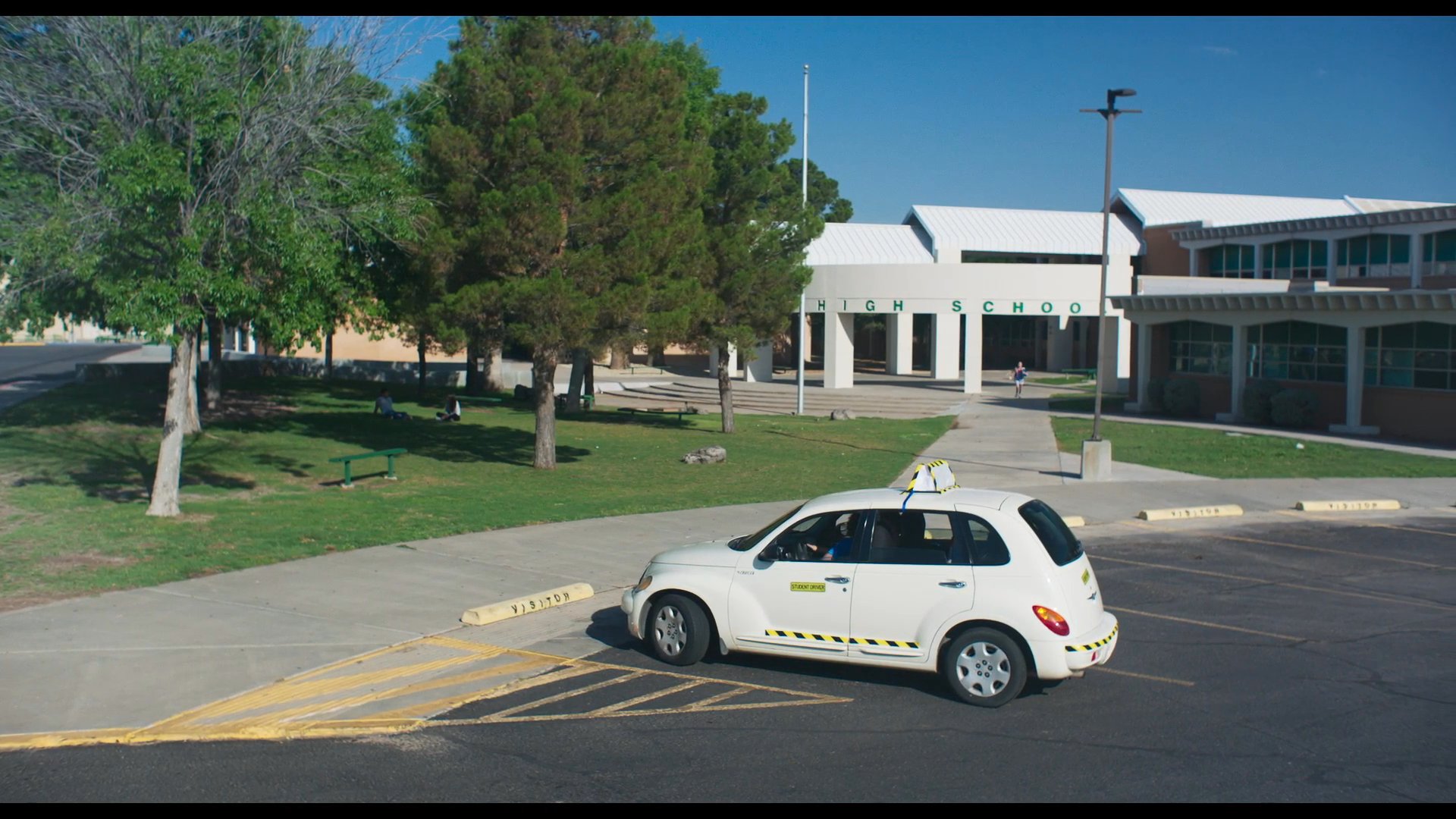 A student driver maneuvers a vehicle through a school parking lot while an instructor provides guidance. After the car comes to a stop, a student exits the building and runs toward the vehicle.