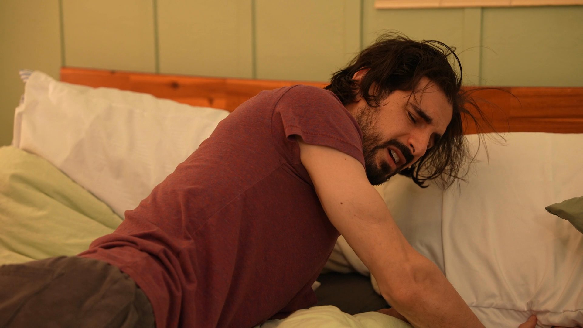 A man in visible emotional distress lies on a bed while a therapist observes and engages with him in a home-based care setting.