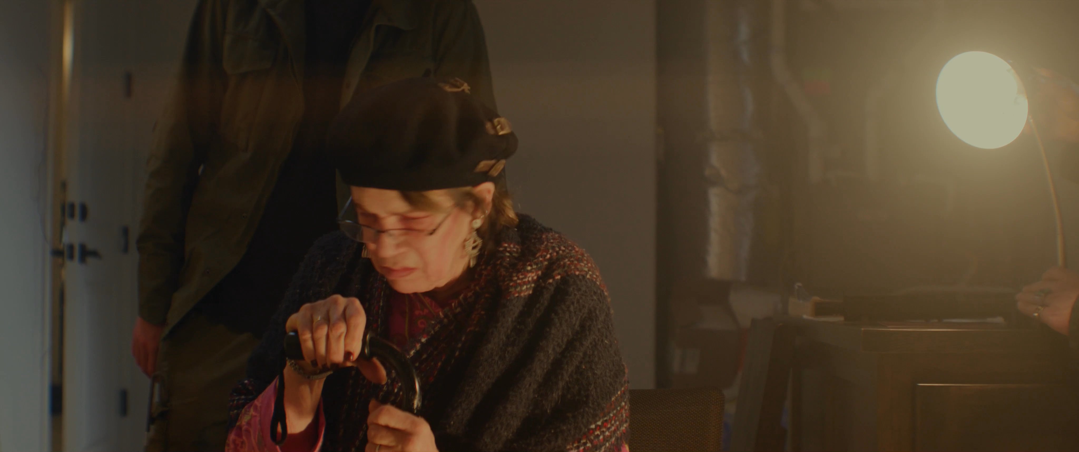 Individuals engage in a serious, low-light conversation within an indoor setting, with characters exchanging dialogue and interacting near a desk lamp.