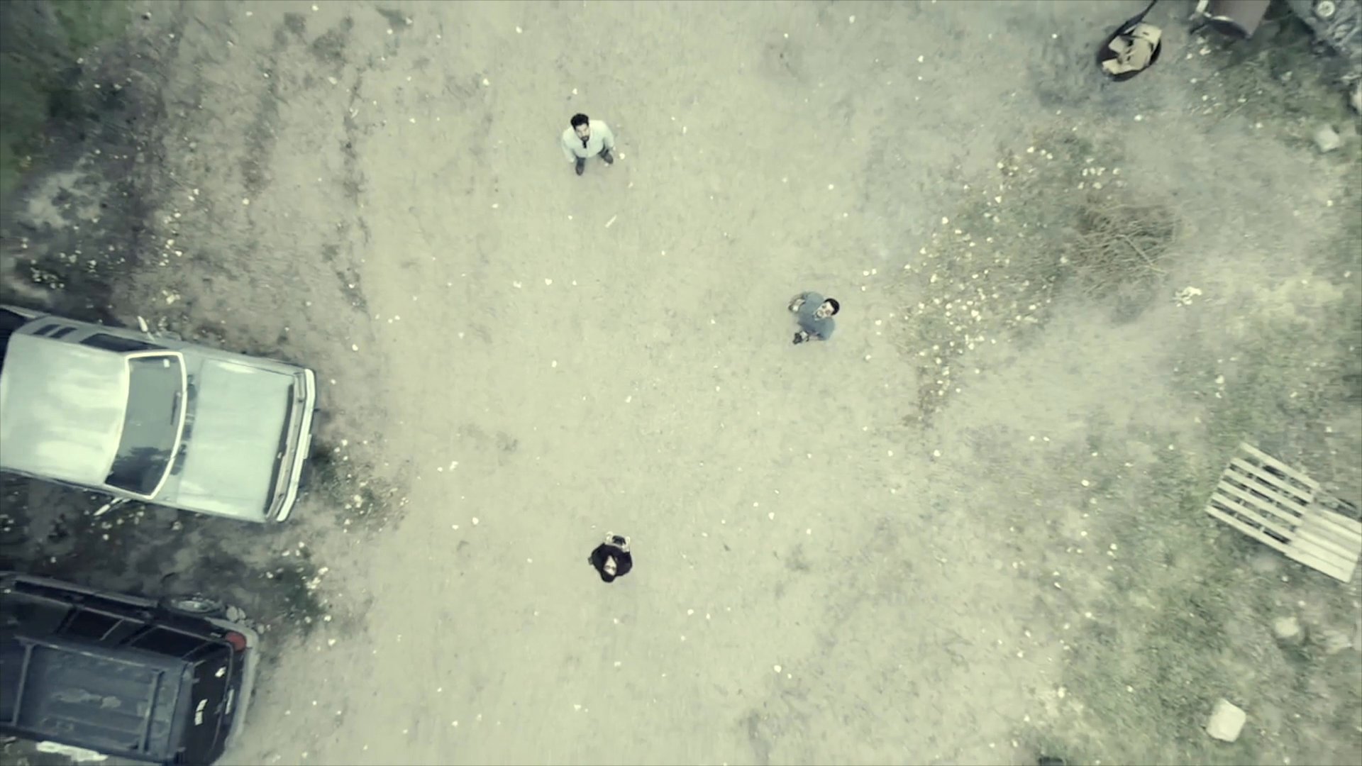A high-angle drone shot observes multiple individuals standing in a desolate, dirt-covered clearing one month after a tragedy. The camera slowly ascends, distancing the subjects from the camera's view as they remain positioned in the open area.