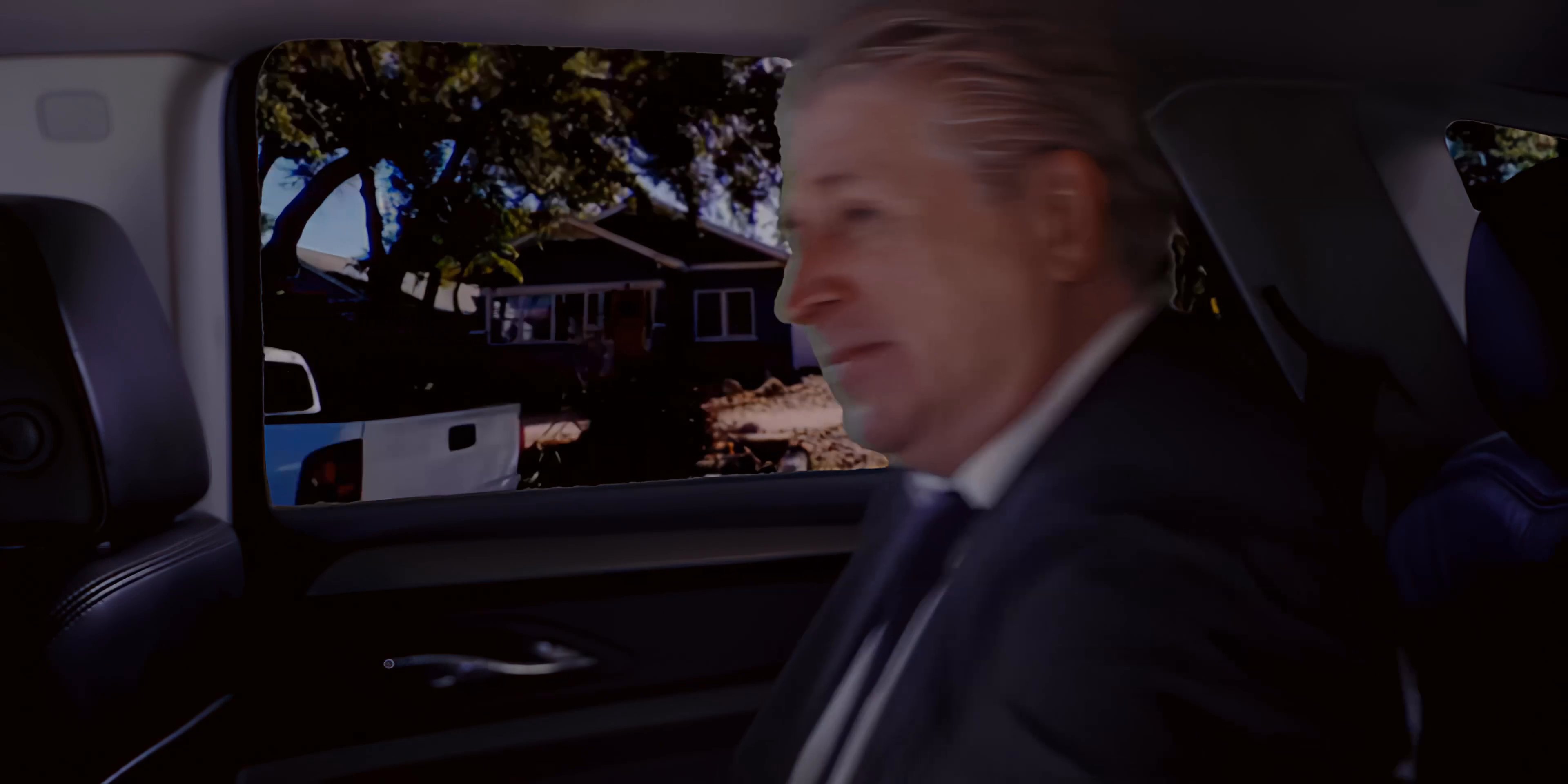 Two men travel in the backseat of a vehicle while discussing business, transitioning to aerial perspectives of a city skyline and highway interchange.