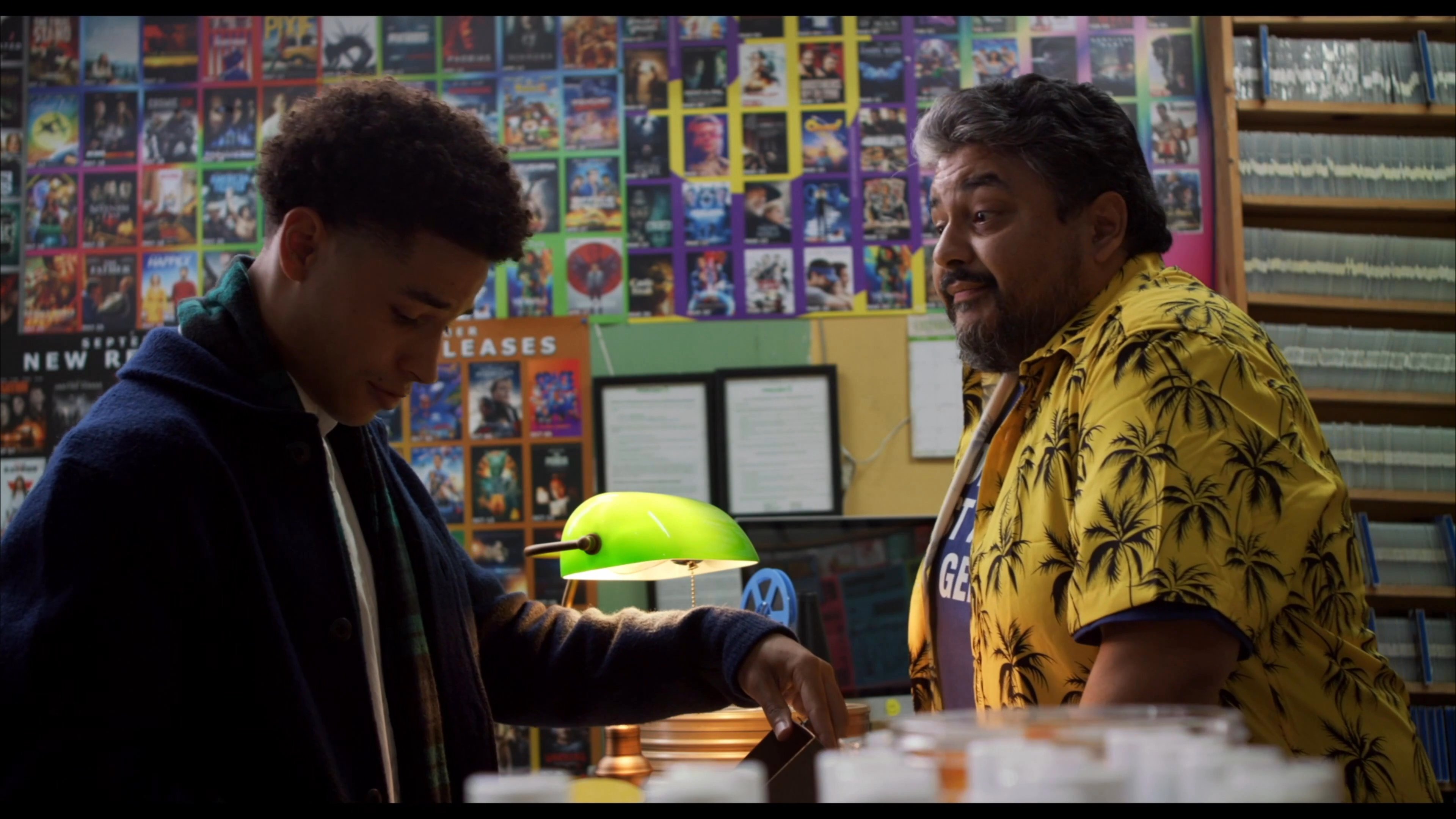 A customer interacts with a store clerk in a vintage video rental shop while discussing a collection of film reels. The dialogue captures a casual yet professional exchange between the two individuals in a retail setting.