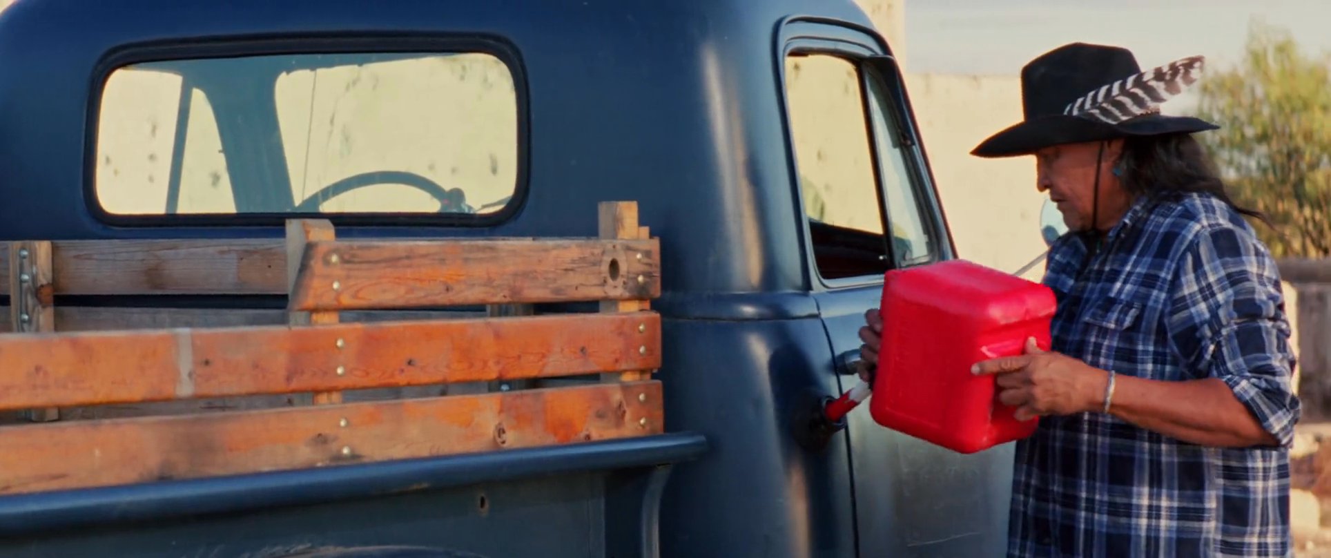 A man in a cowboy hat refills a vintage pickup truck with fuel while horses stand nearby in a rural landscape. A second man interacts with him as they work together on the vehicle.